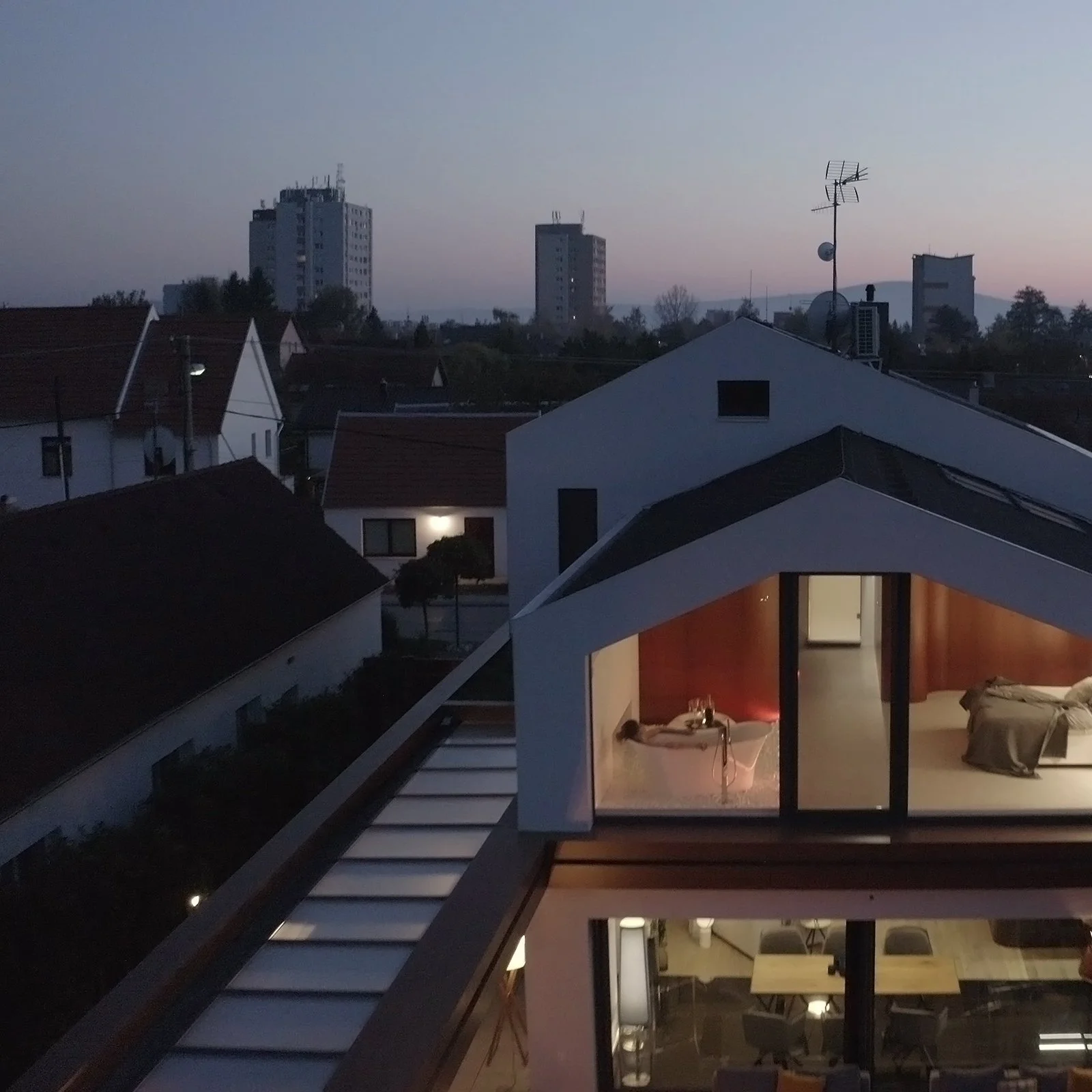 A rooftop view at dusk showing neighboring houses, some taller buildings in the distance, and a bedroom with a person lying on a bed visible through large windows.