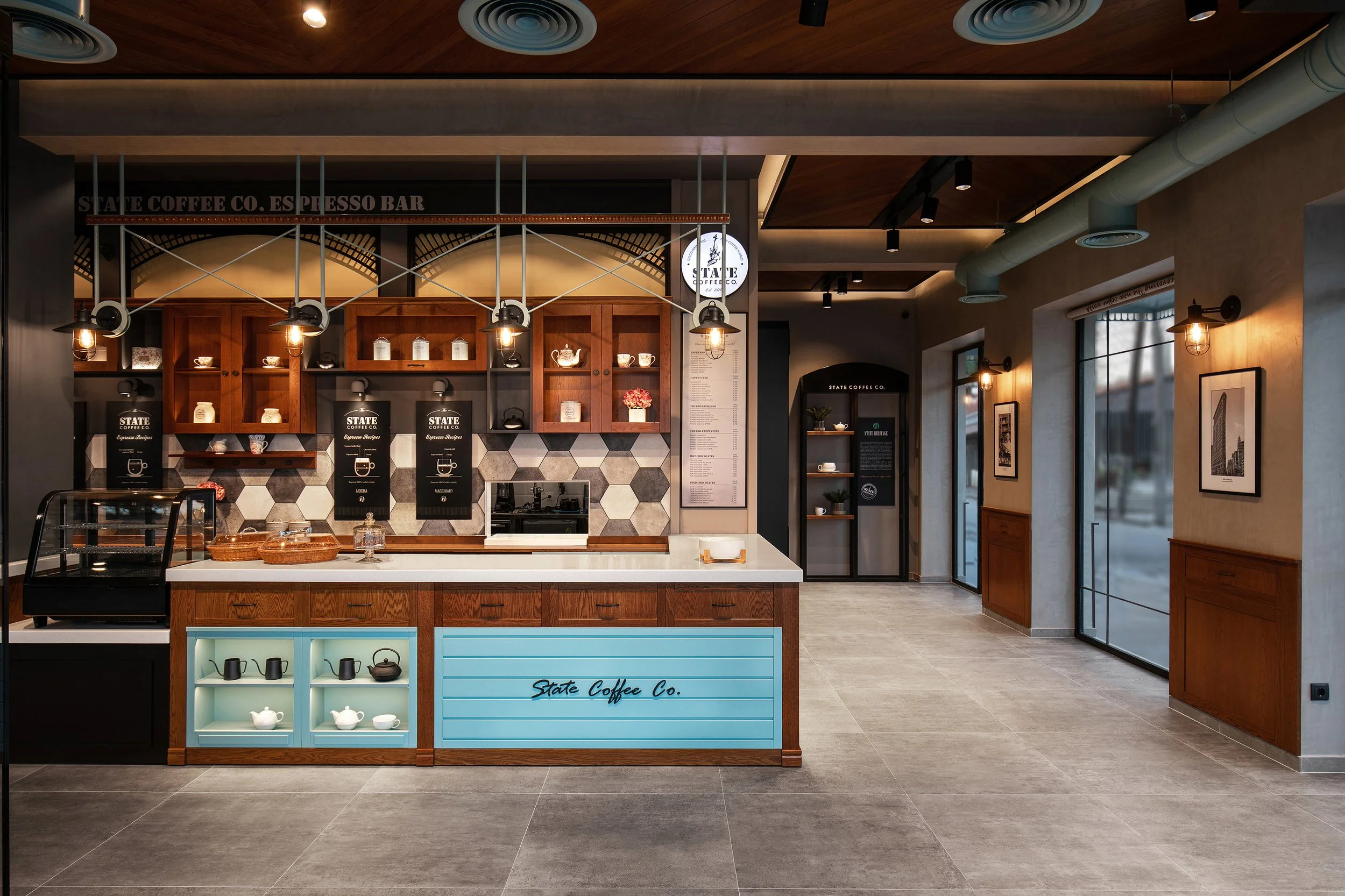 Interior of a modern coffee shop with a wooden counter, black and white patterned backsplash, hanging lights, and framed artwork on the walls.