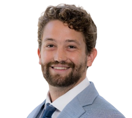 A man with curly hair and a beard, smiling, wearing a light blue suit and white shirt, against a plain white background.