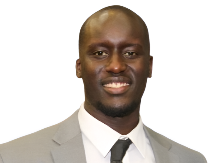 Portrait of a smiling man with a dark complexion, wearing a light gray suit, white shirt, and black tie, against a plain white background.