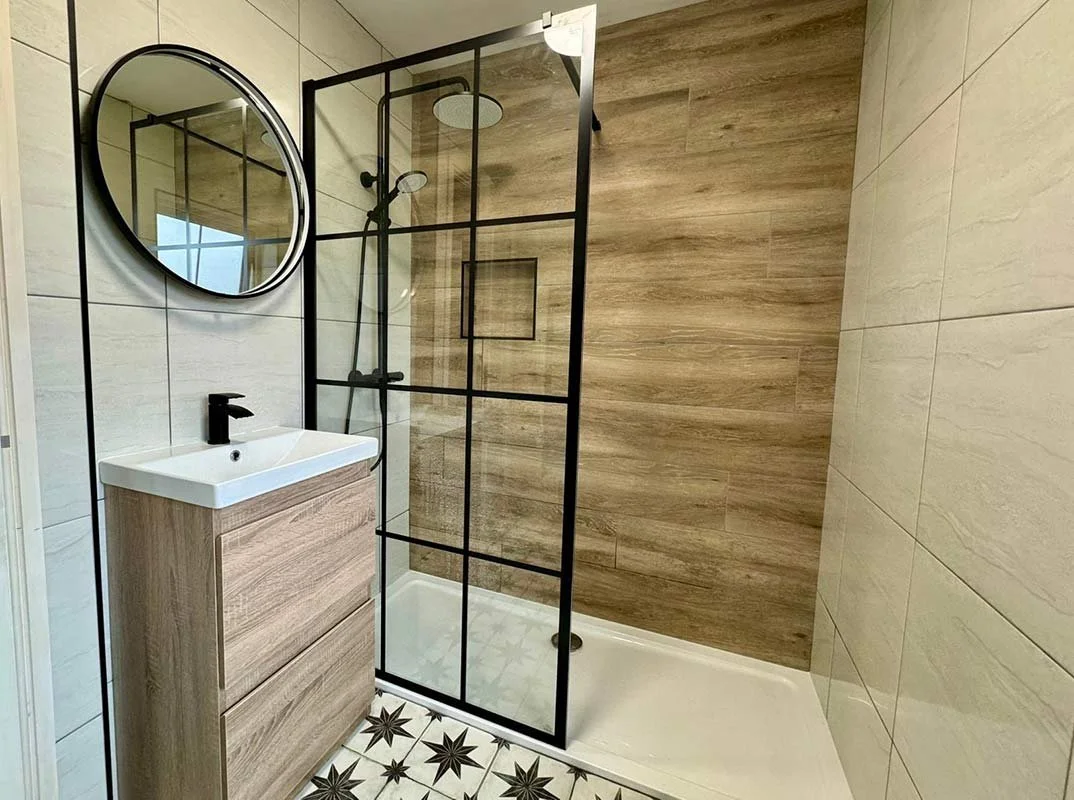 Modern bathroom with a shower area enclosed by a black metal frame, a round mirror, a small white sink with wood cabinet, beige tiles, and star-patterned floor tiles.