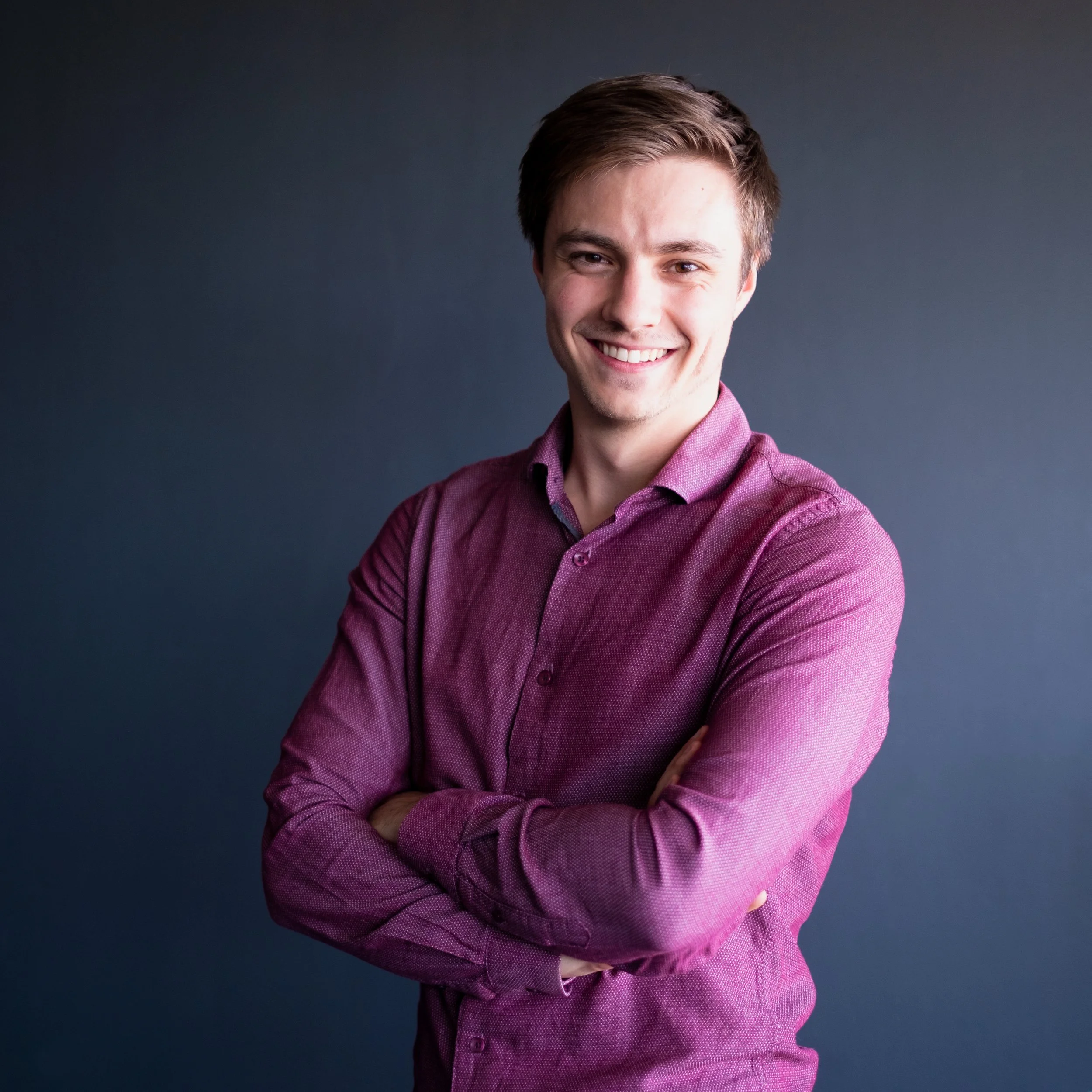 Jeune homme souriant en chemise bordeaux avec les bras croisés contre un fond gris.