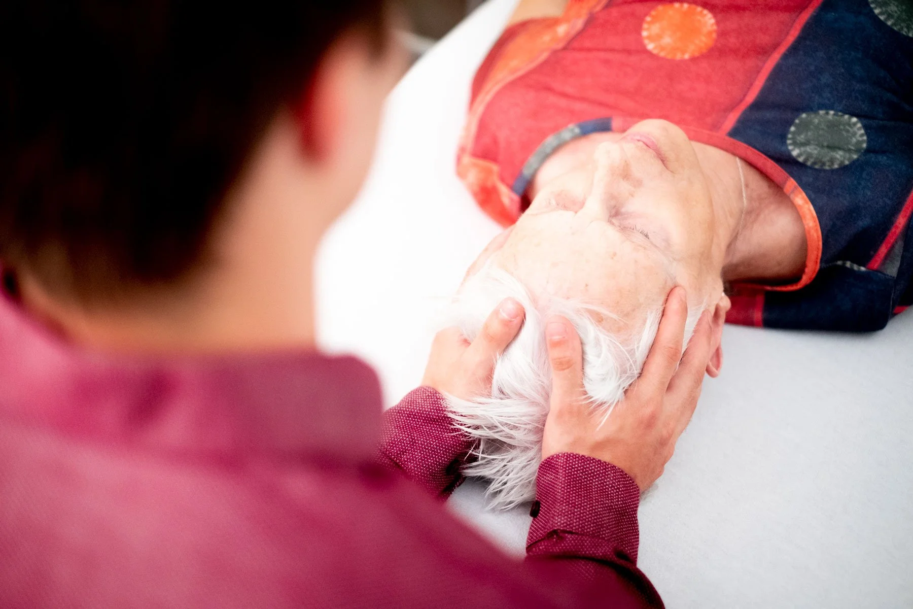 Une personne âgée reçoit un soin de la part d'une autre personne, un ostéopathe, qui tient sa tête dans un contexte de soins ou de réconfort.