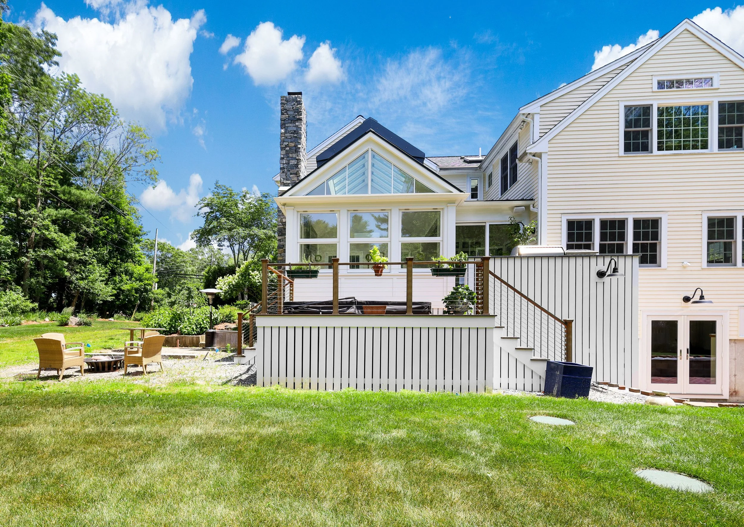Backyard with a large house featuring a deck with plants, outdoor seating, and a grassy lawn under a bright blue sky.