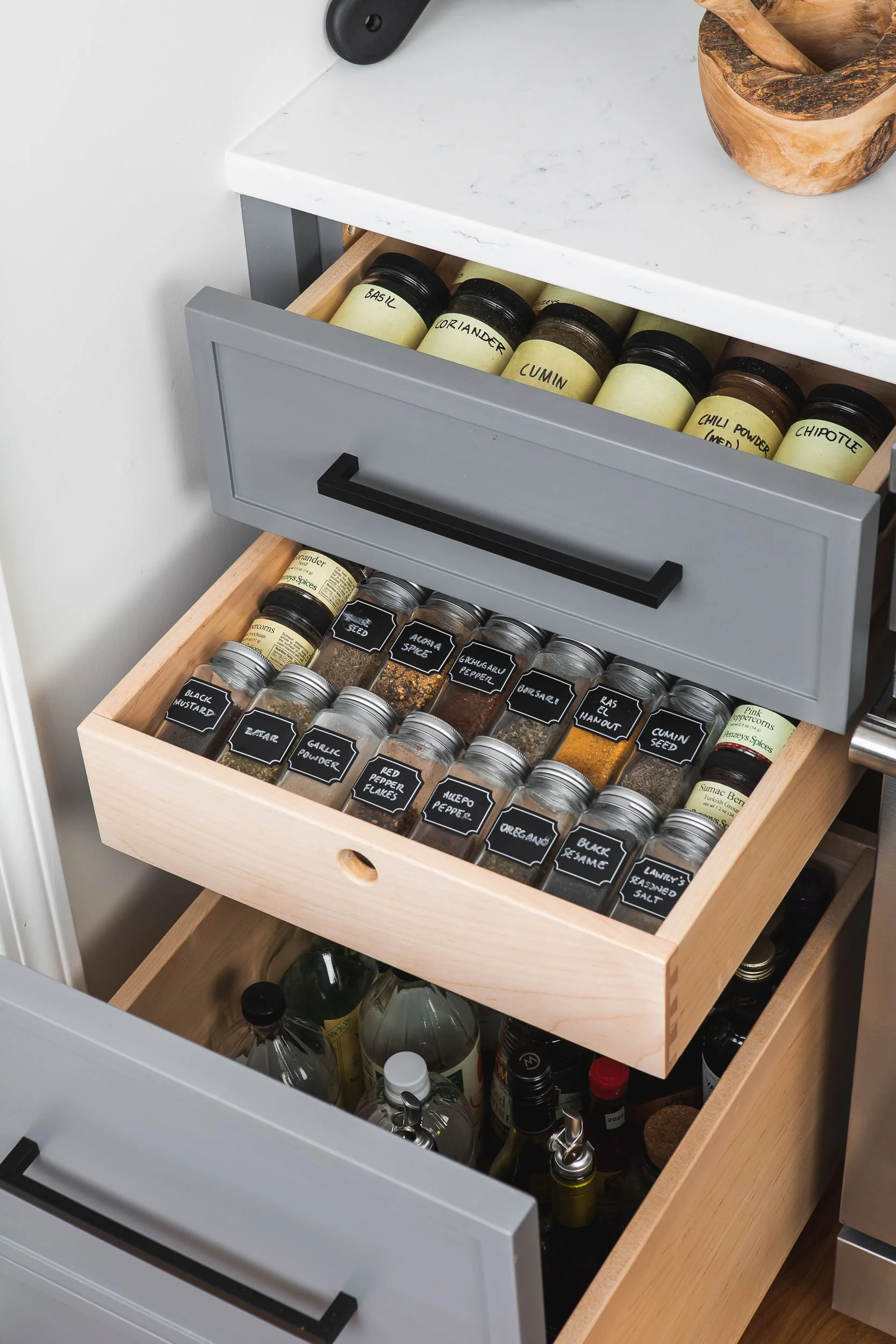 Open kitchen drawer with spices and seasonings in small glass jars, labeled with black chalkboard tags, including garlic powder, black mustard, cumin seed, and oregano.
