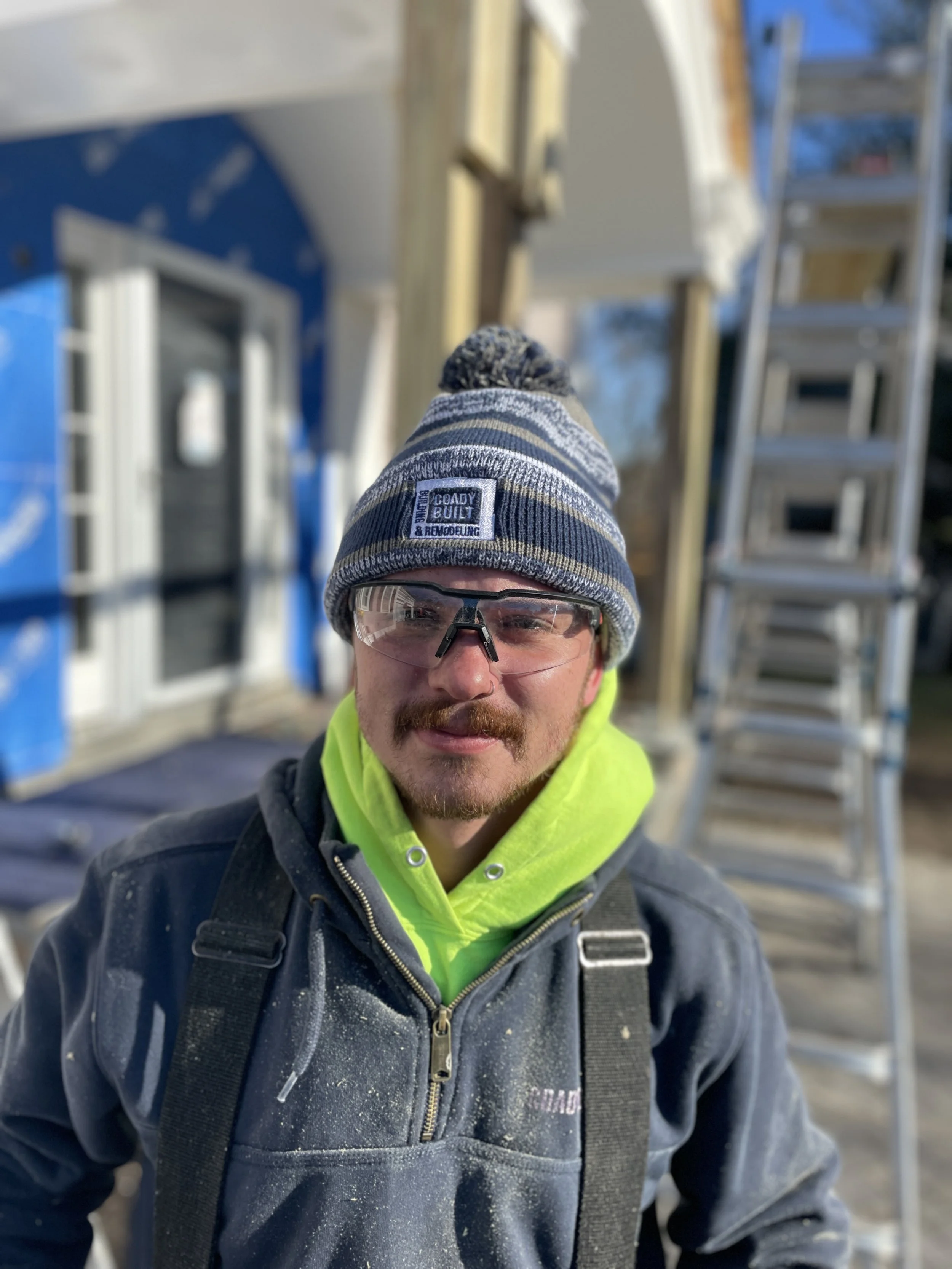Scott Cargill, Coady Built's Carpenter, wearing a beanie, safety glasses, a neon yellow hoodie, and a dark jacket with construction dust, standing in front of a building under construction with a ladder.