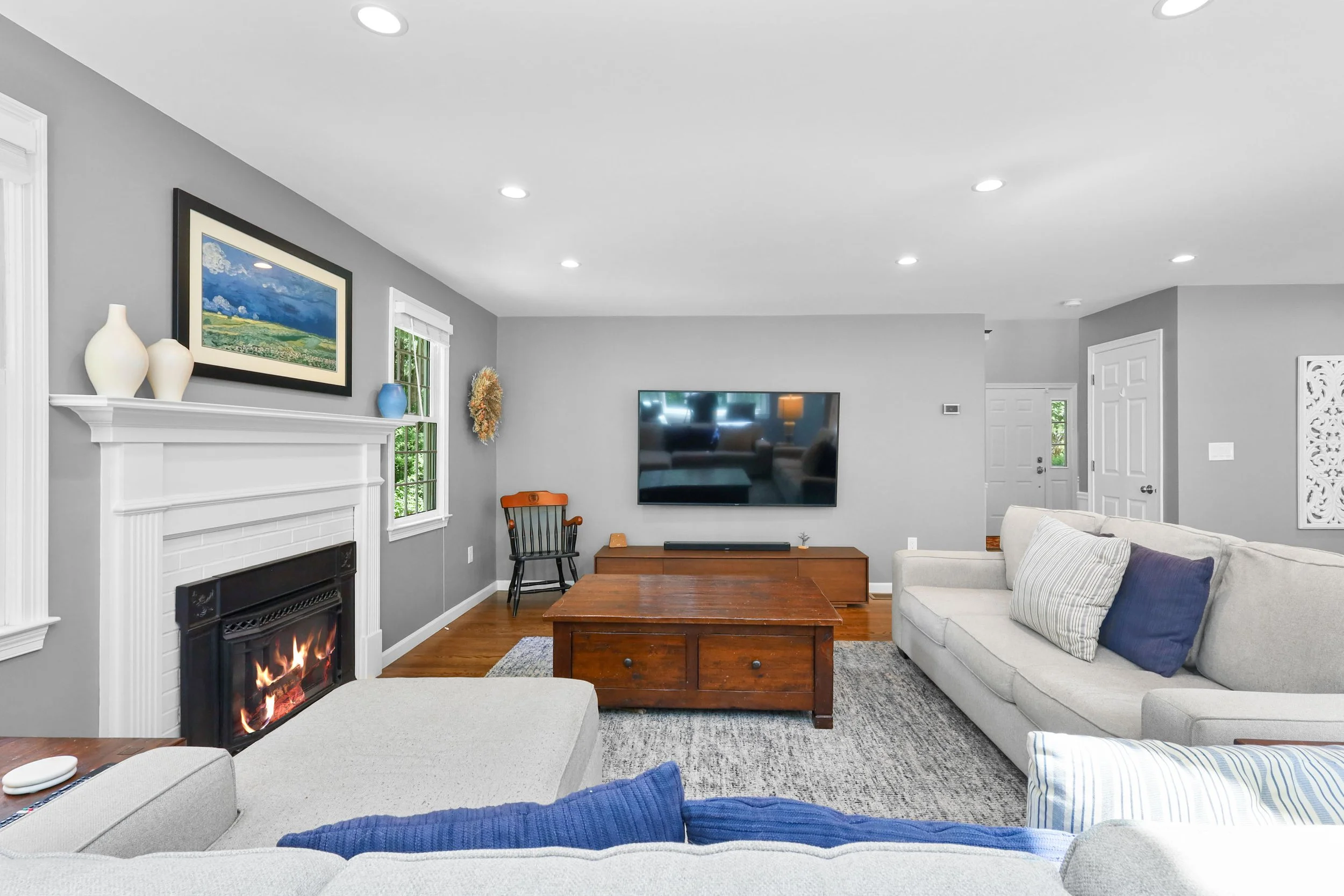 Living room with gray walls, white fireplace with decorative vases, three windows, mounted flat-screen TV, wooden coffee table, and two sofas with cushions, decorative carpet, and colorful wall art.