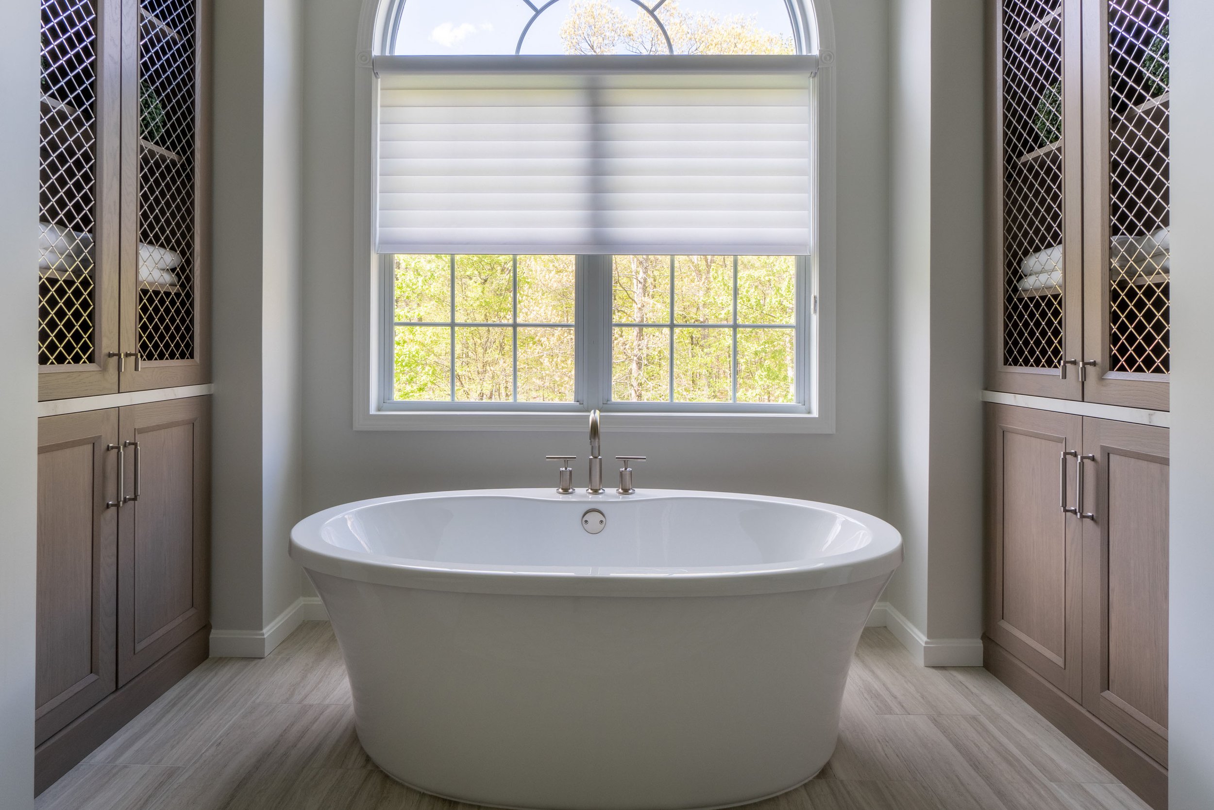 Modern bathroom with white freestanding bathtub beneath a large window with a white roller shade, flanked by built-in cupboards with lattice doors, and light wood flooring.