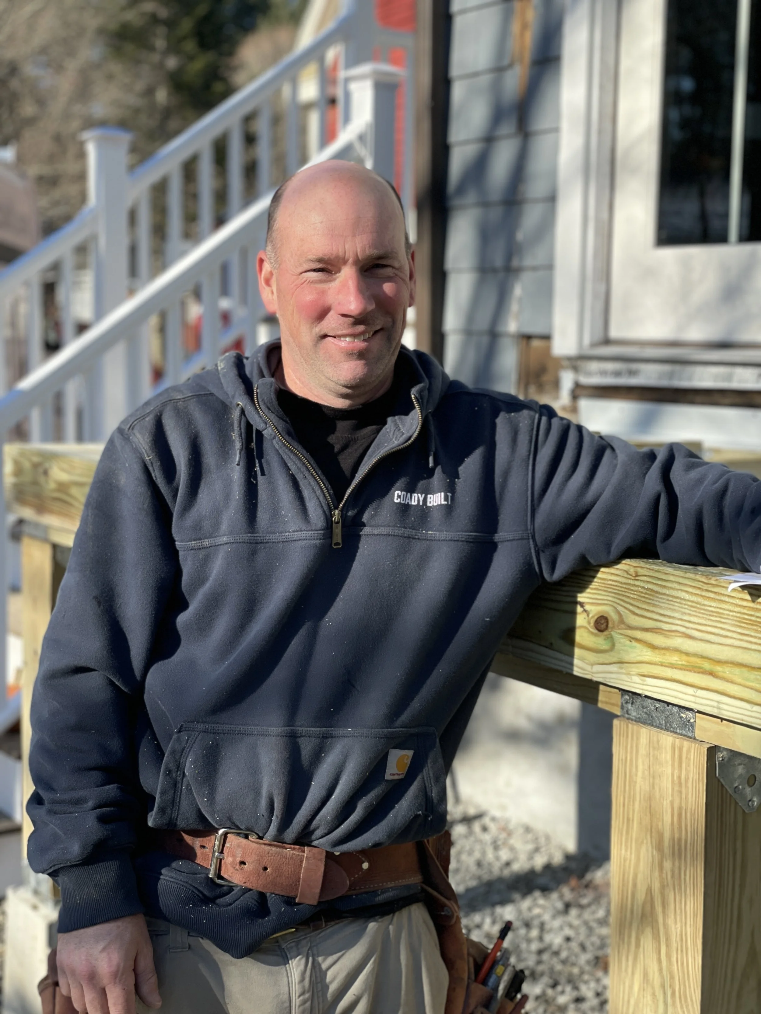 Jeremy Melcher, Lead Carpenter and Construction Manager at Coady Built, smiling on a construction site, wearing a black Carhartt hoodie, with a wooden railing and house under construction in the background.