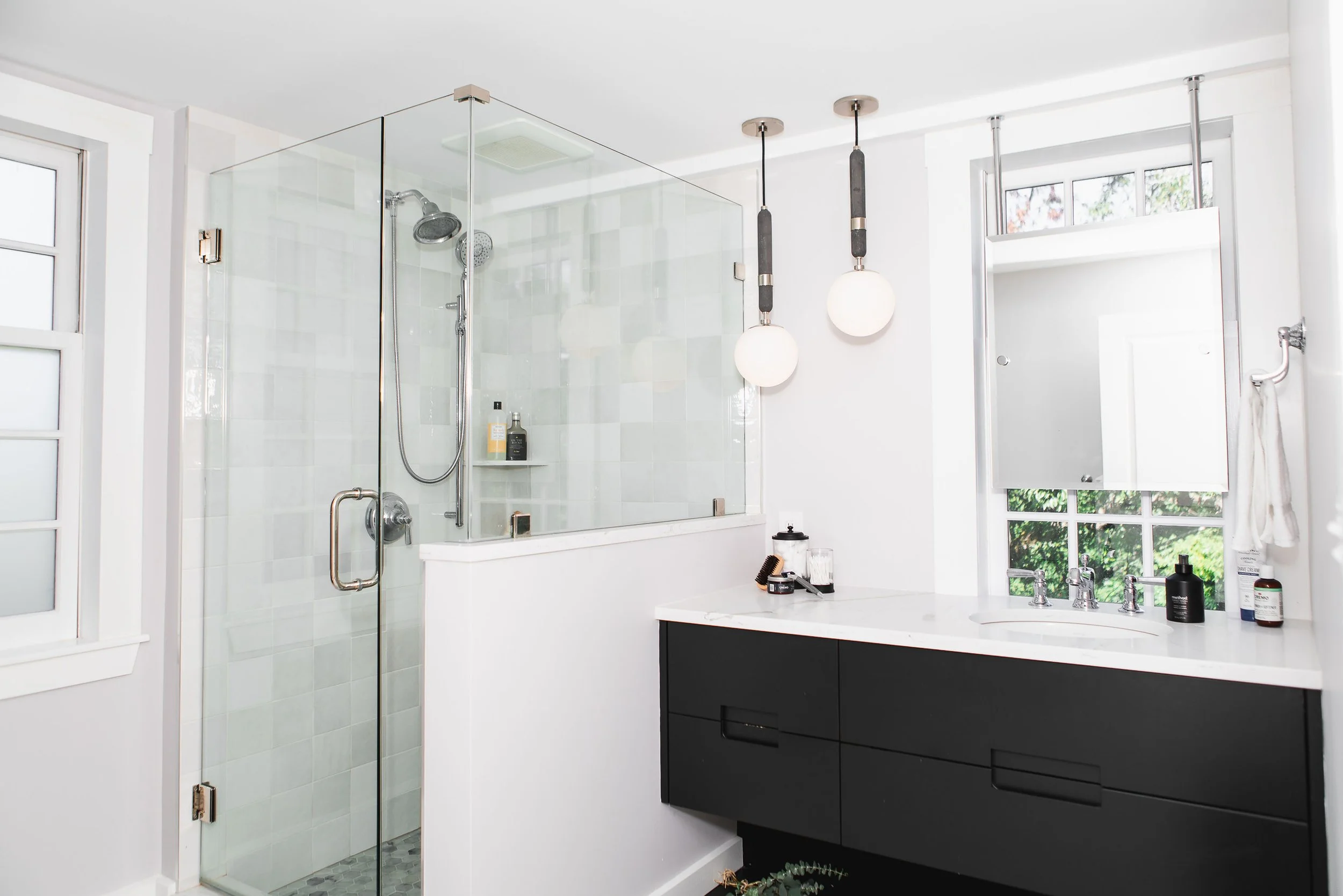 Modern bathroom with a glass shower enclosure, black vanity, white countertop, dual sinks, mirror, hanging light fixtures, and window with greenery outside.
