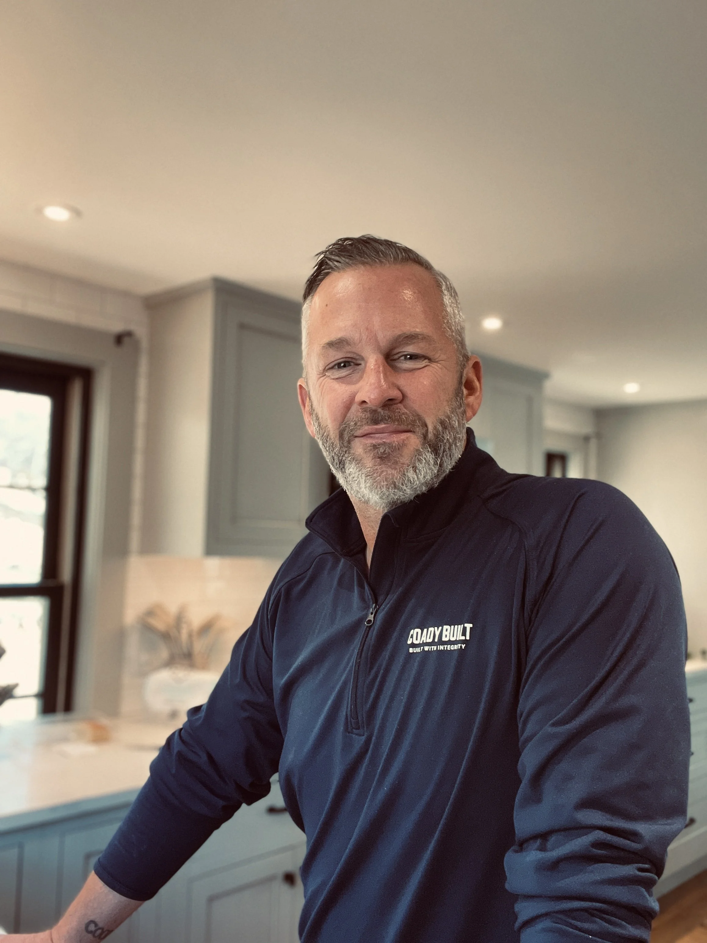 Matt Coady, Founder of Coady Built, wearing a navy blue jacket with 'COADY BUILT' logo, standing in a modern kitchen with gray cabinets and a window.