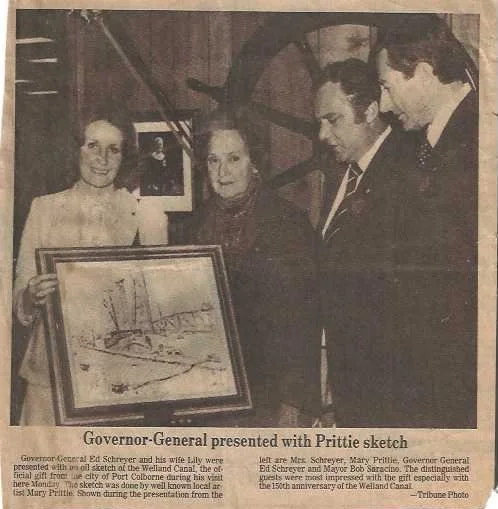 Four people standing and holding a framed sketch of ships and a river in an indoor setting, with a fifth person standing on a staircase in the background.