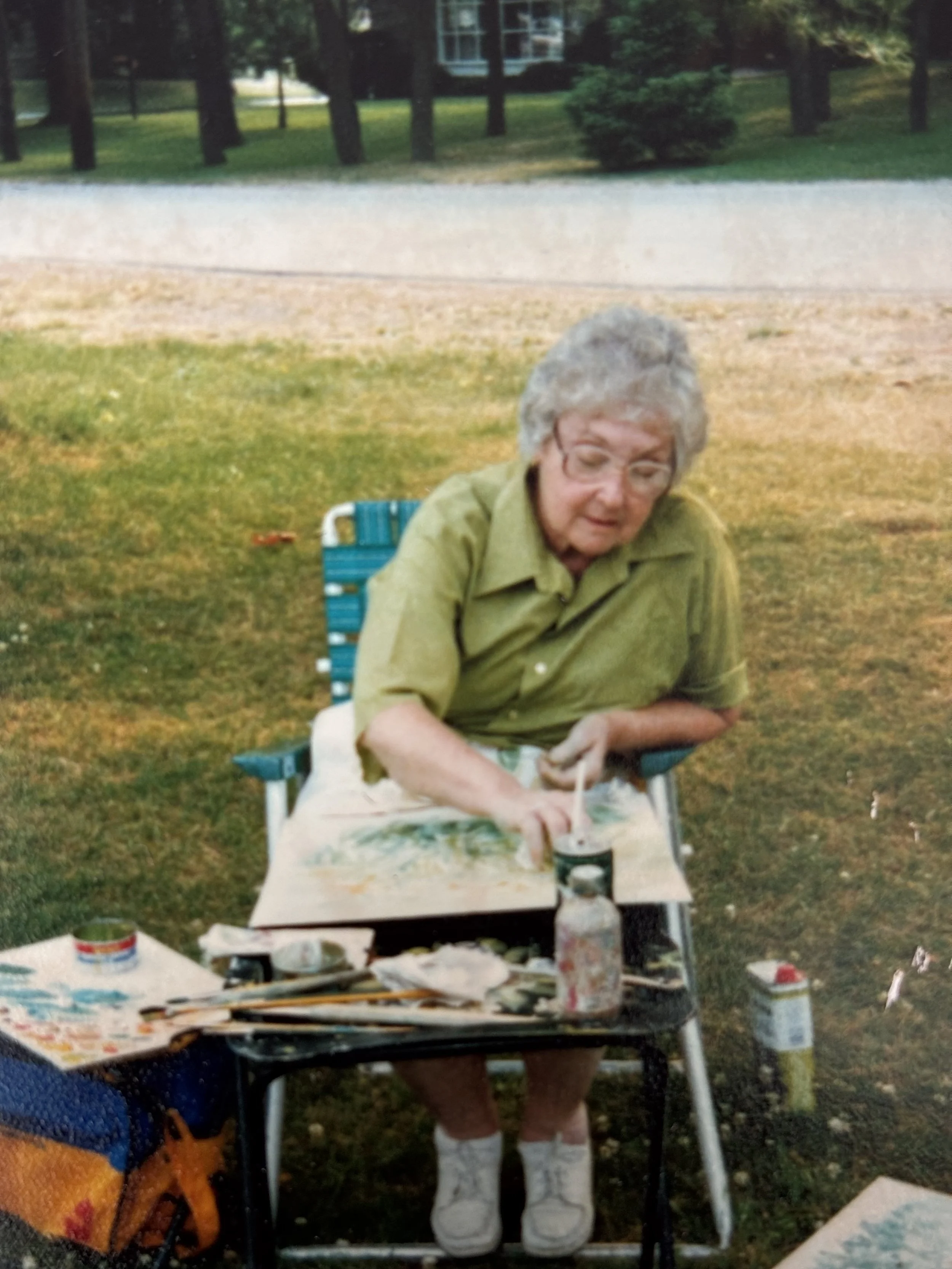 Mary Prittie sitting in a folding lawn chair outdoors, painting on a canvas set on a small table in front of her.  She is wearing a green shirt, white socks, and white shoes, with art supplies and paint jars on the table.