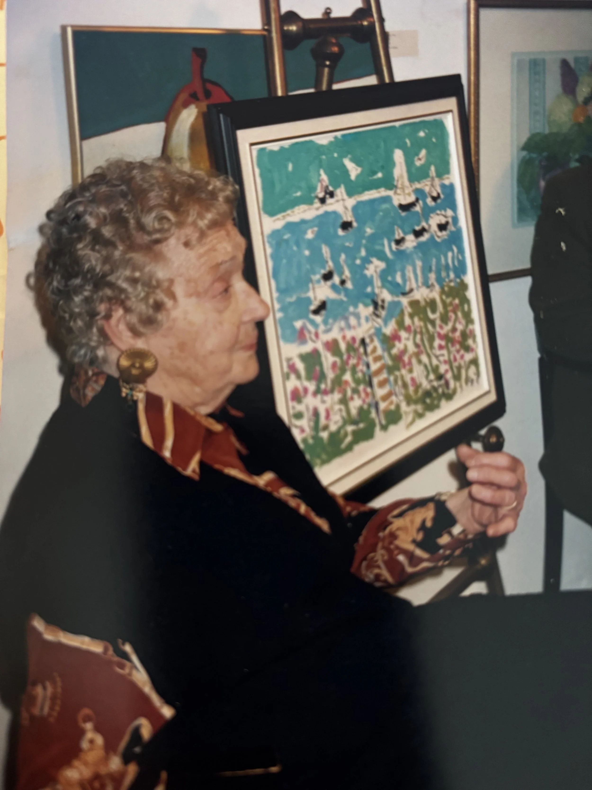 Mary Prittie, wearing a dark outfit and large gold earrings, is holding and looking at a colorful painting of sailboats on water, set against a vibrant background with trees and pink flowers.
