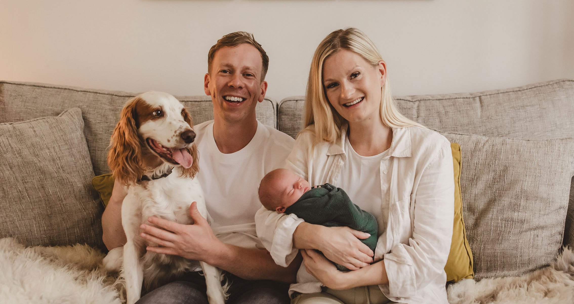 A happy family with a dog, a newborn baby, and a woman sitting on a beige sofa in a cozy living room.