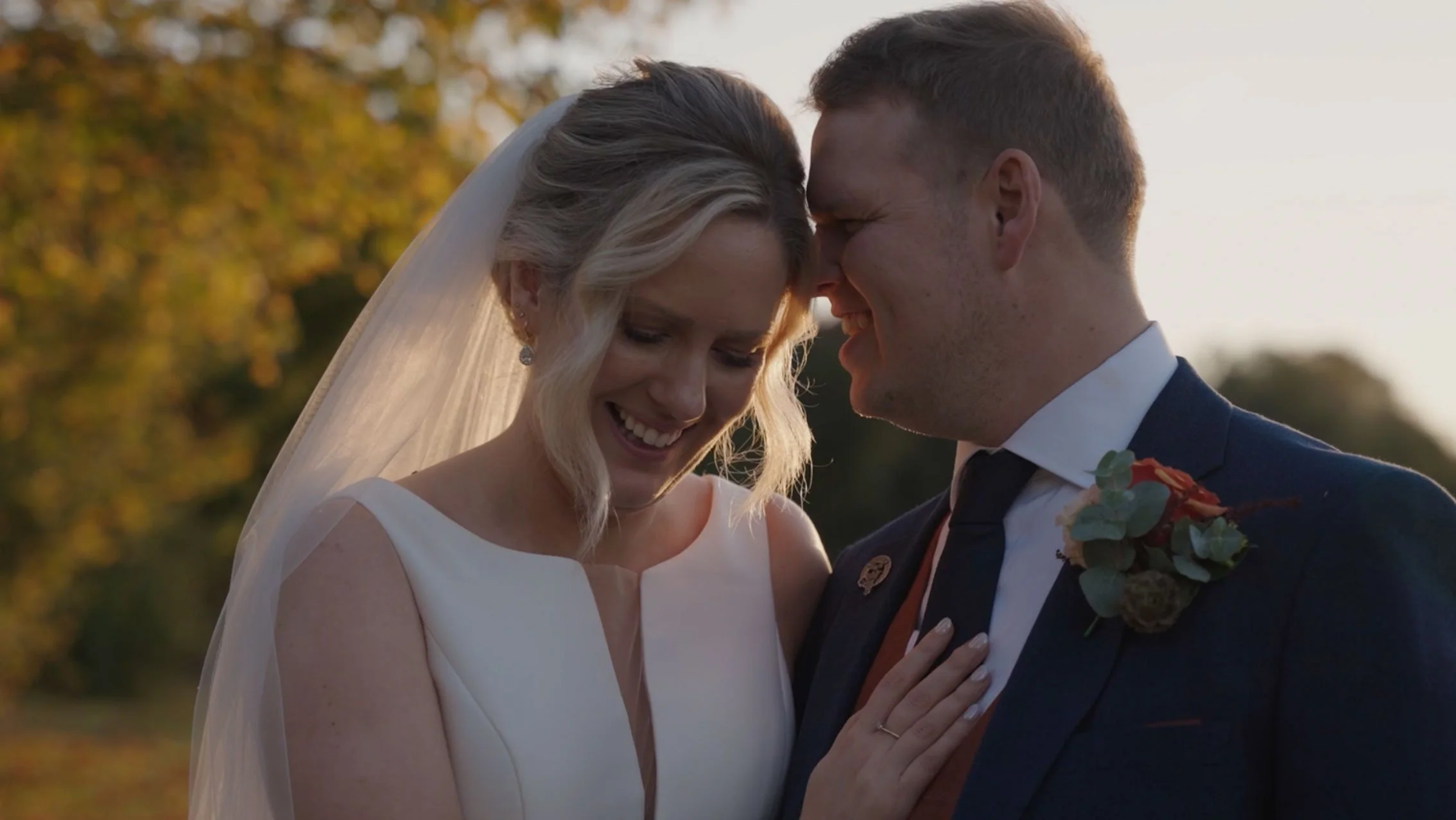 A bride and groom smiling with their foreheads touching outdoors during sunset, with autumn trees in the background.