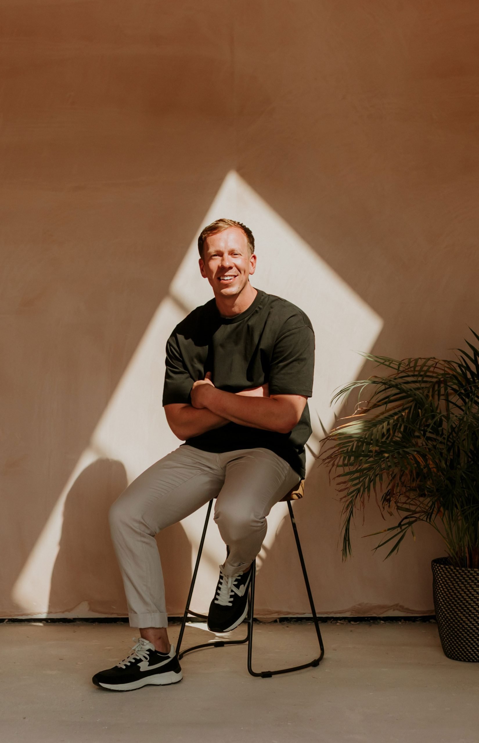 A smiling man with short hair, wearing a black t-shirt, beige pants, and black sneakers, sitting on a black metal chair and crossing his arms, in front of a wall with a large white diamond-shaped shadow and a potted plant.