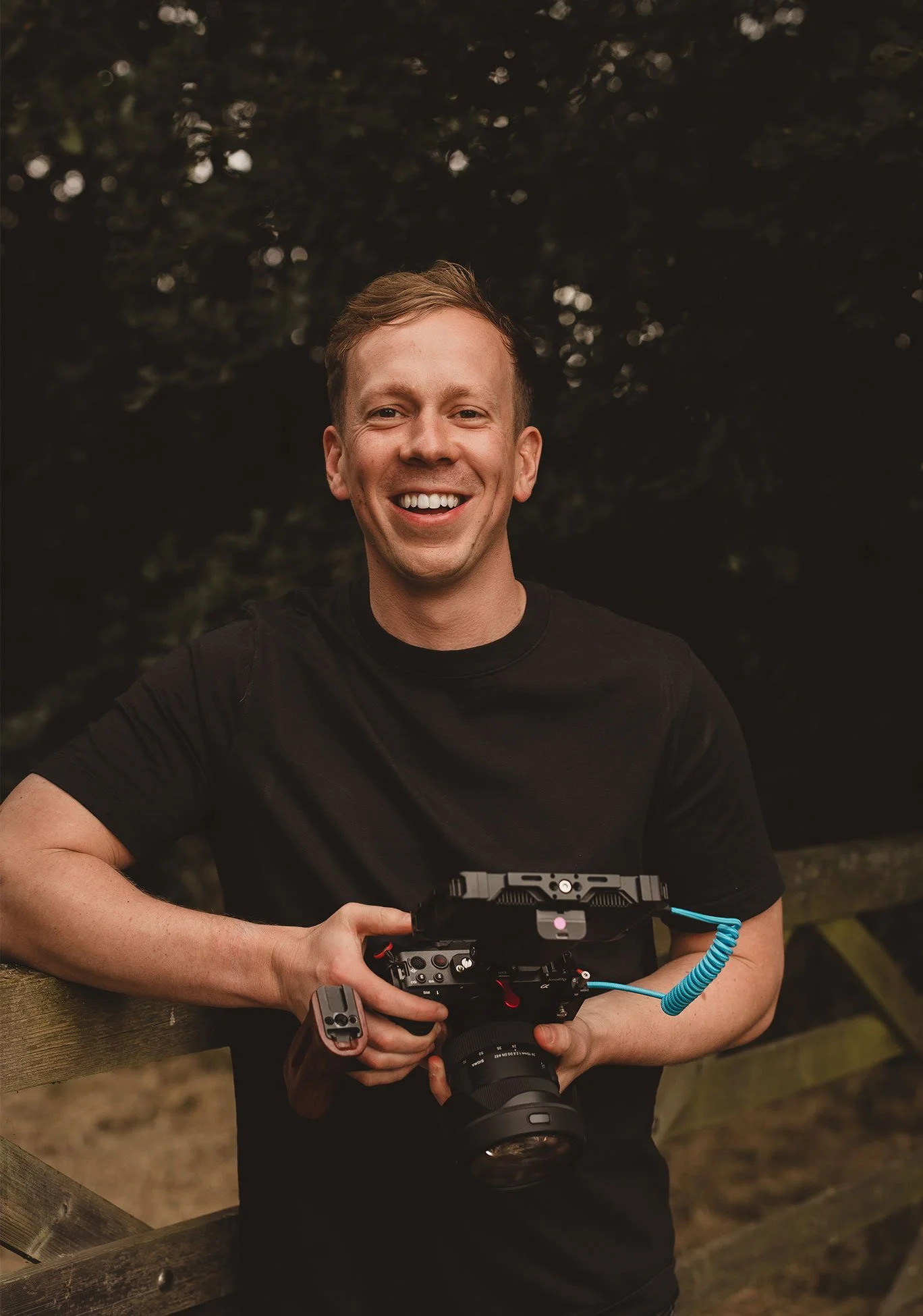 A smiling man in a black shirt holding a professional camera outdoors, with trees in the background.