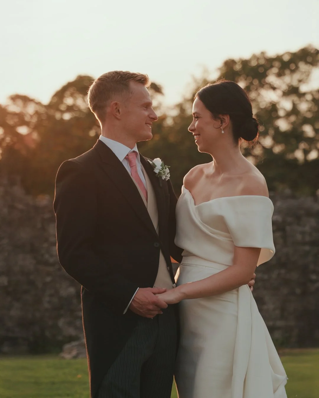 A newlywed couple stands close together, smiling at each other, with a sunset and trees in the background. The groom wears a black suit with a pink tie and boutonniere, and the bride wears an off-shoulder white wedding dress.