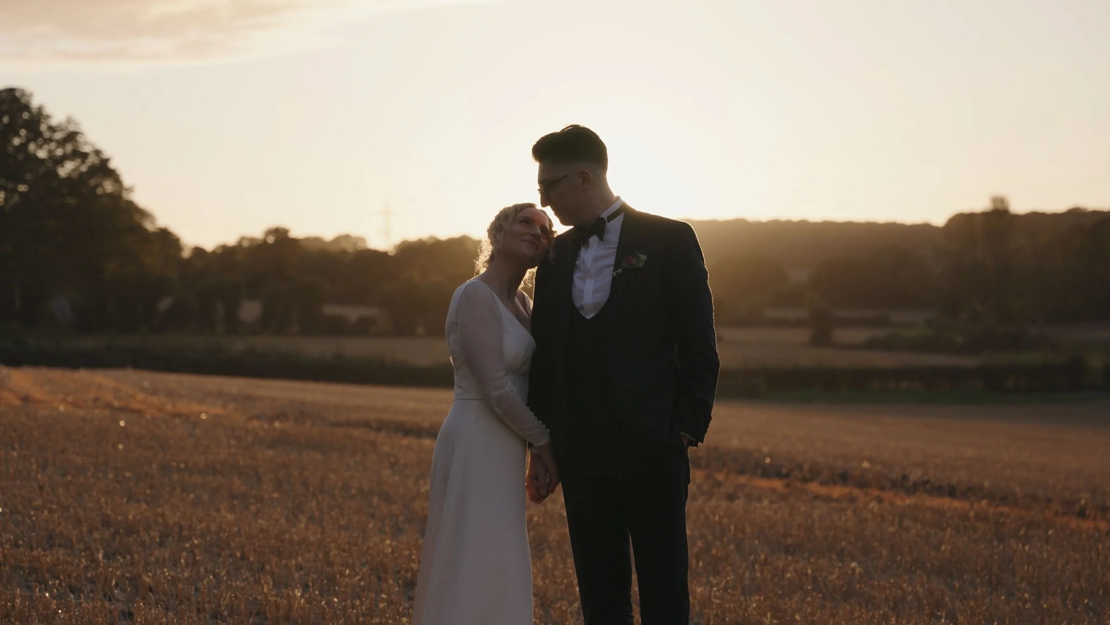 A couple in wedding attire holding hands and standing outdoors at sunset, with the woman resting her head on the man's shoulder.