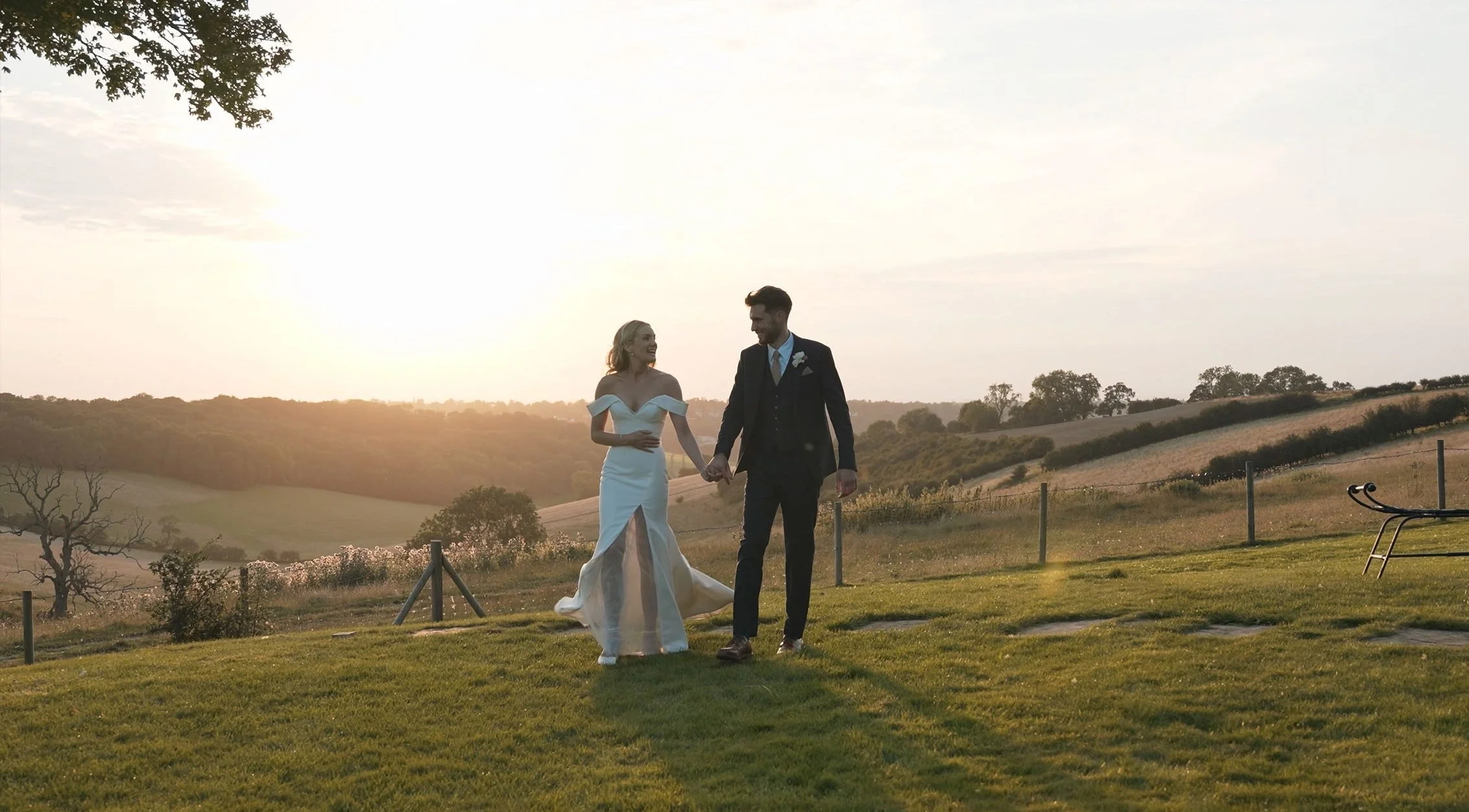 A bride and groom are walking hand-in-hand on a grassy outdoor area during sunset, with rolling hills and trees in the background.