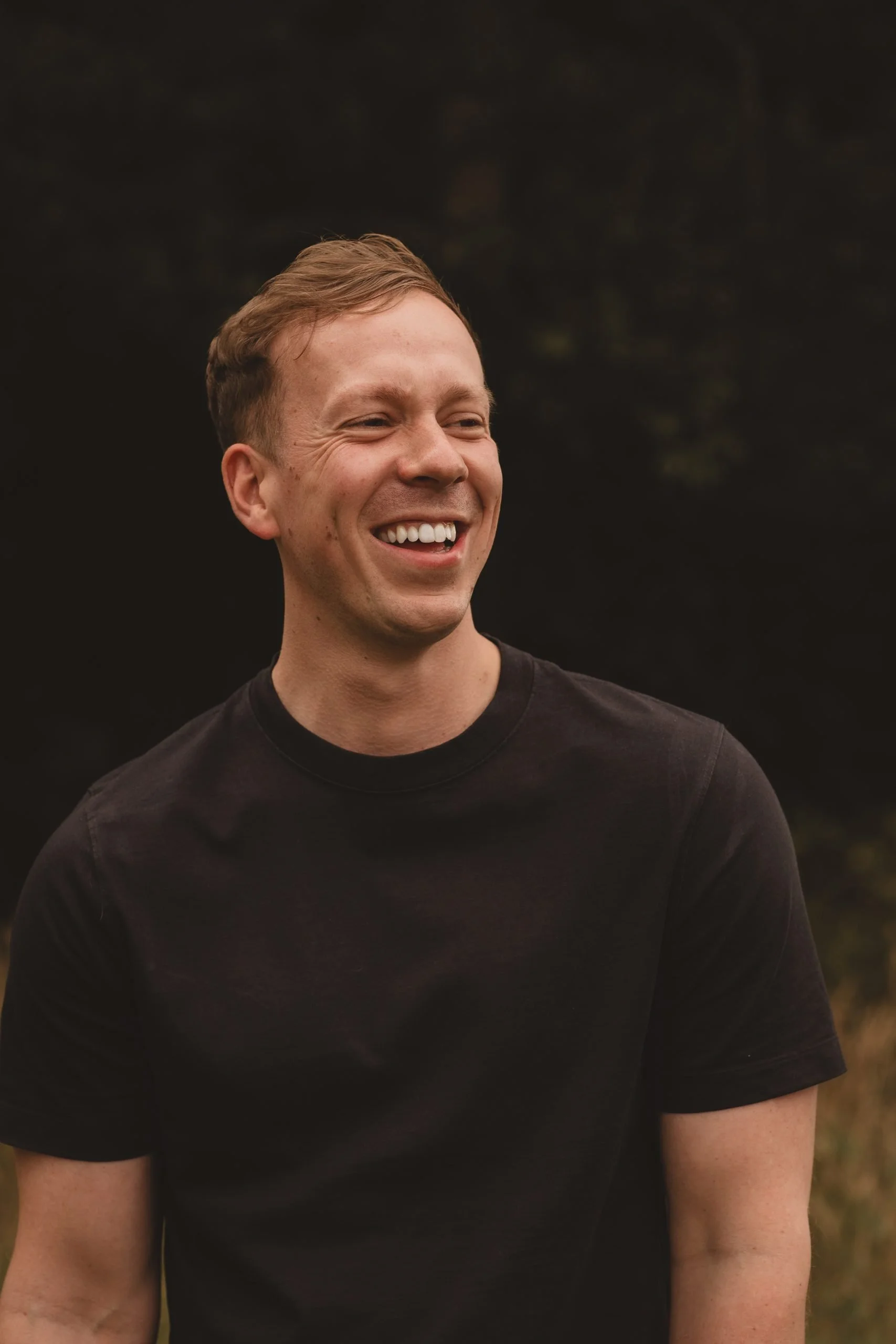 A young man with short, light brown hair, smiling and laughing outdoors against a dark, blurred background, wearing a black t-shirt.