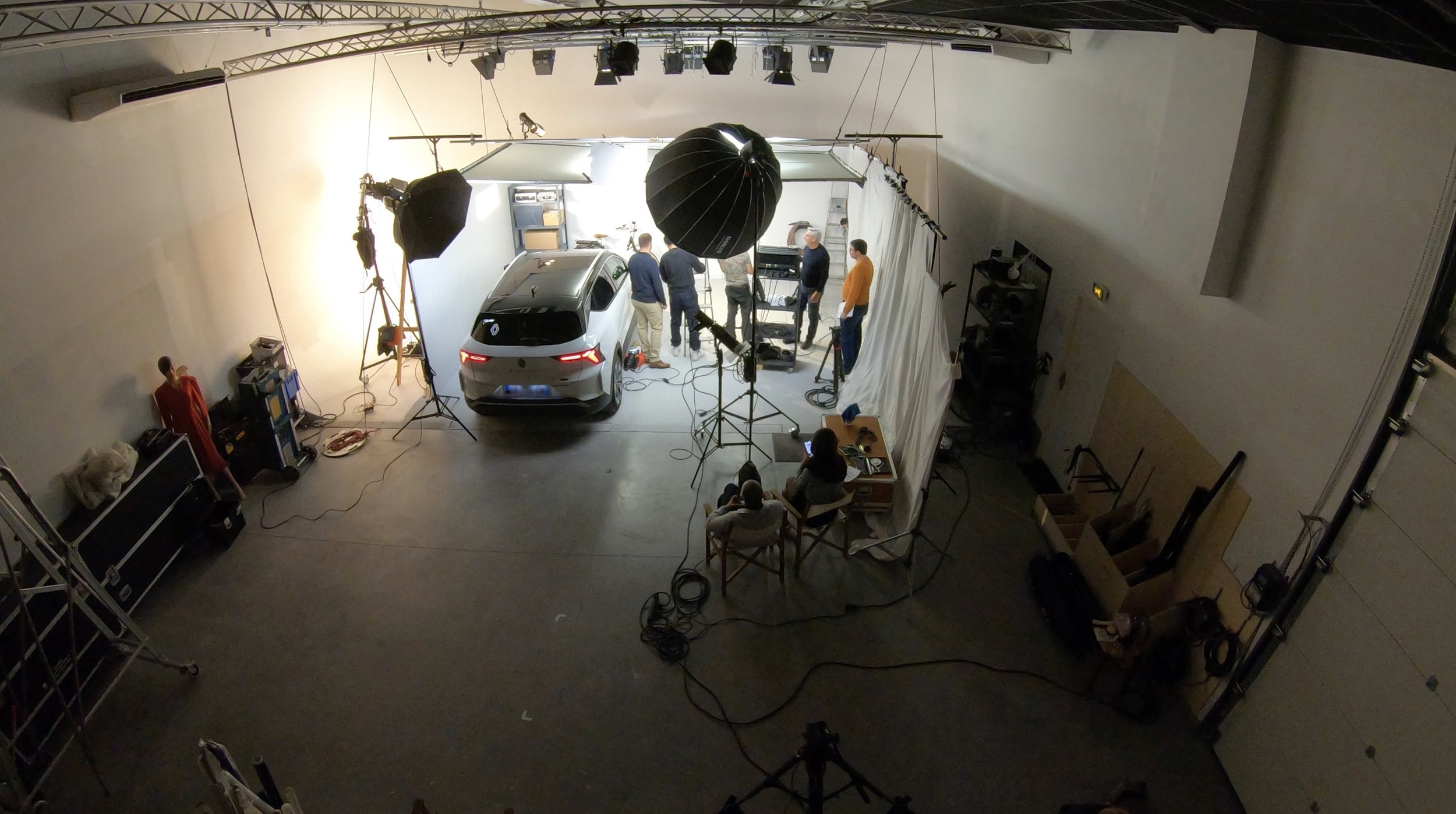 Un studio de photographie ou de tournage avec un véhicule (voiture) au centre, entouré d’éclairage professionnel, de câbles, et de plusieurs personnes qui discutent ou travaillent sur un fond blanc. On voit également du matériel de studio, comme des 