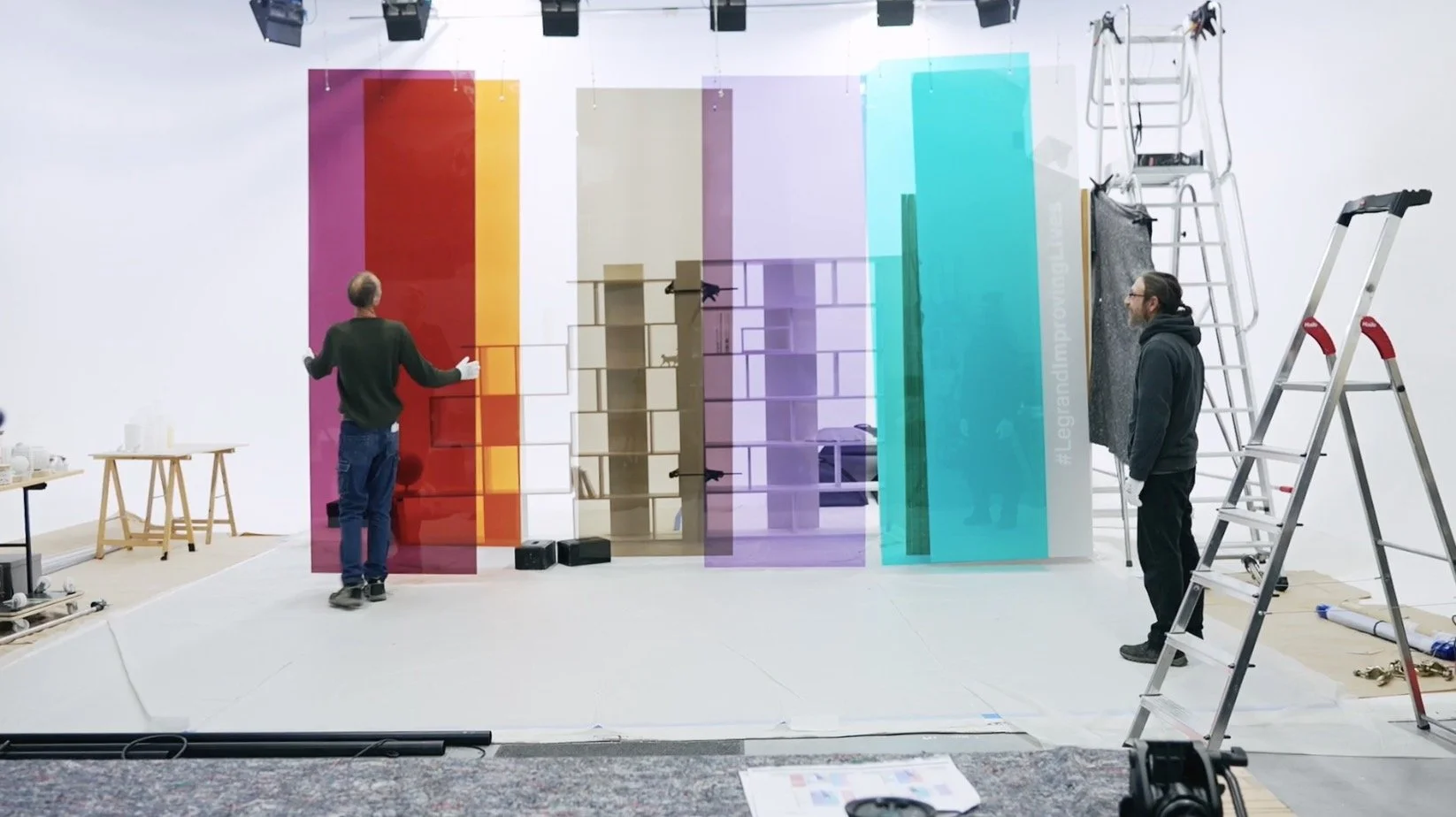 Deux hommes installent une murale colorée dans un studio blanc avec des échelles et des outils.