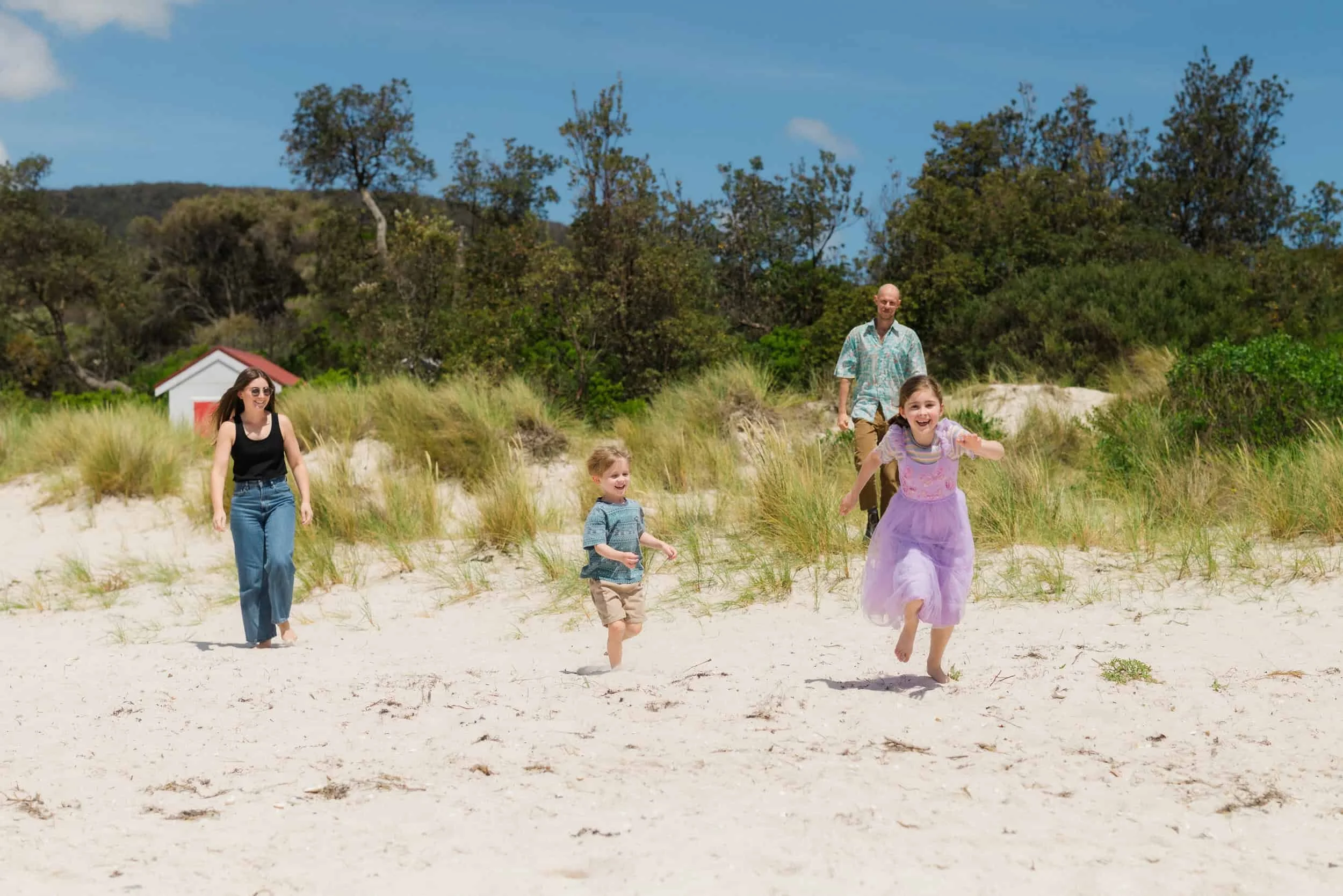 A family of four enjoying a day at the beach, running and playing in the sand with grassy dunes and trees in the background on a sunny day.