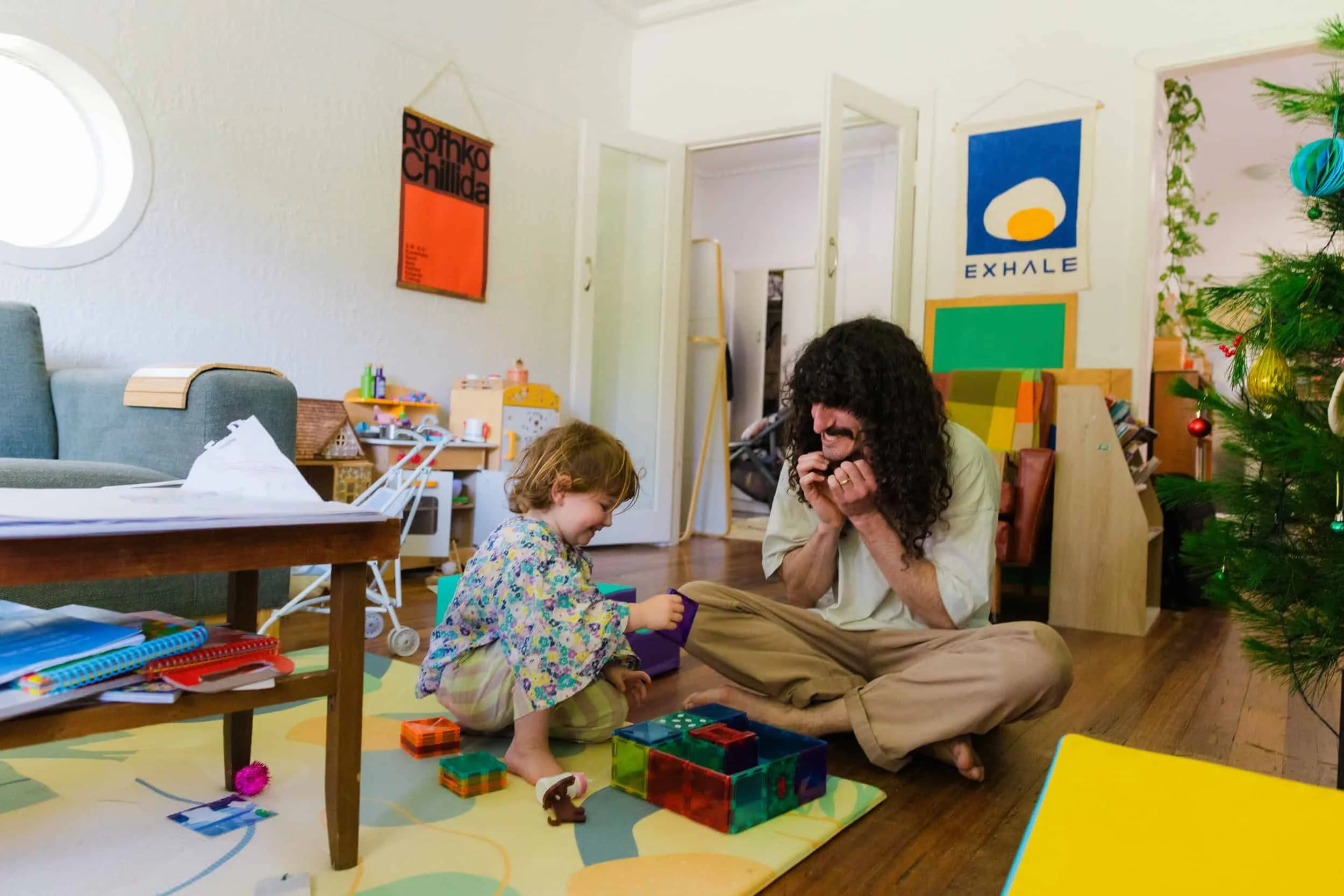 A man with long curly hair and a young girl playing a game on the floor of a colorful, decorated living room. There is a decorated Christmas tree on the right side, with toys and art on the walls.