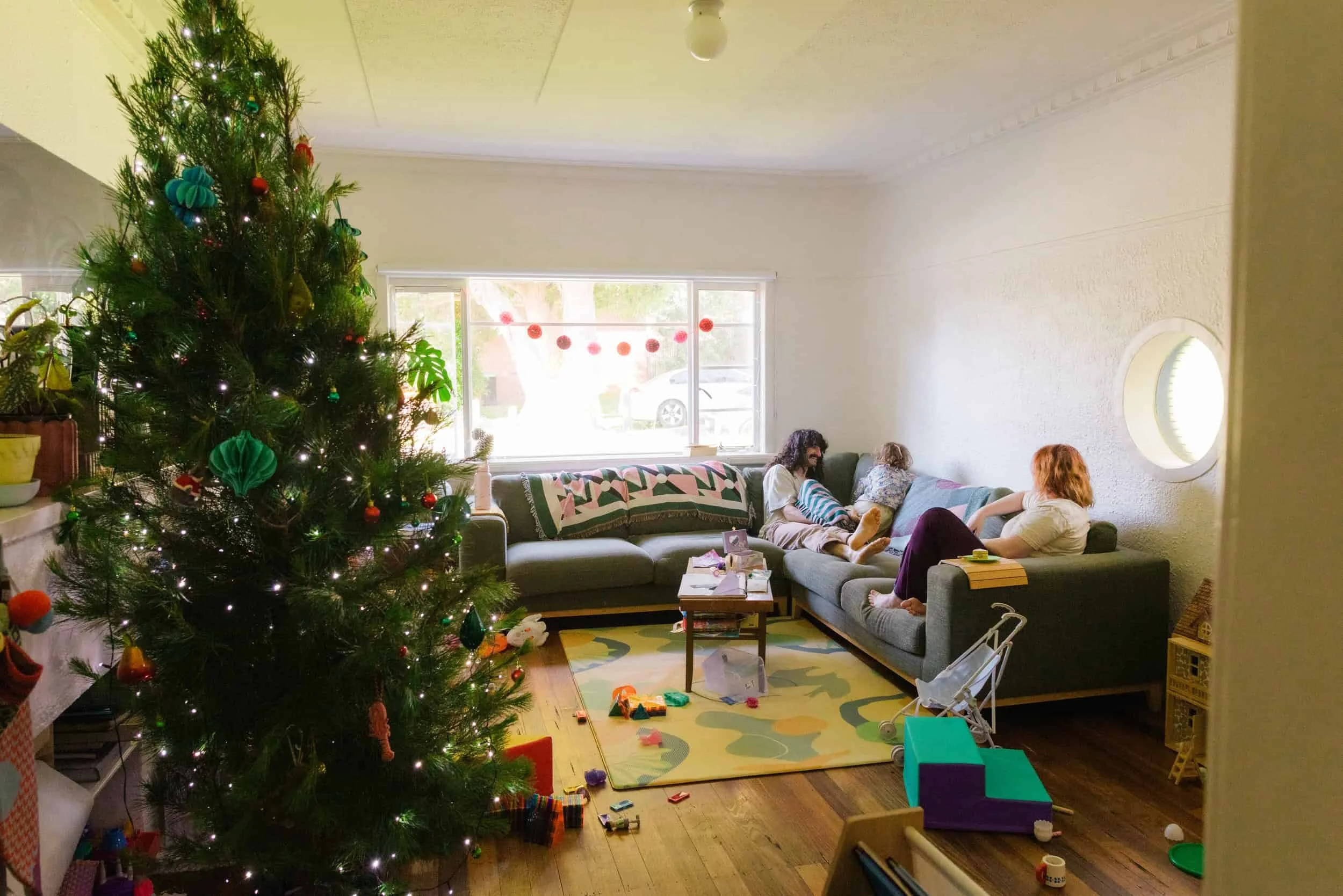 Living room decorated for Christmas with a large Christmas tree, sofa with people, toys scattered on floor, window with red garland.