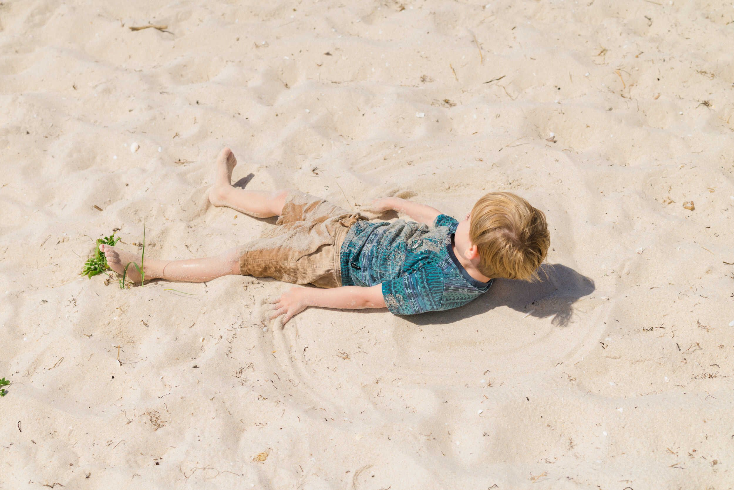 A young boy lying on the sandy beach, facing upward with his head turned to the side, wearing a colorful shirt and khaki shorts, surrounded by small patches of green plants and footprints in the sand.