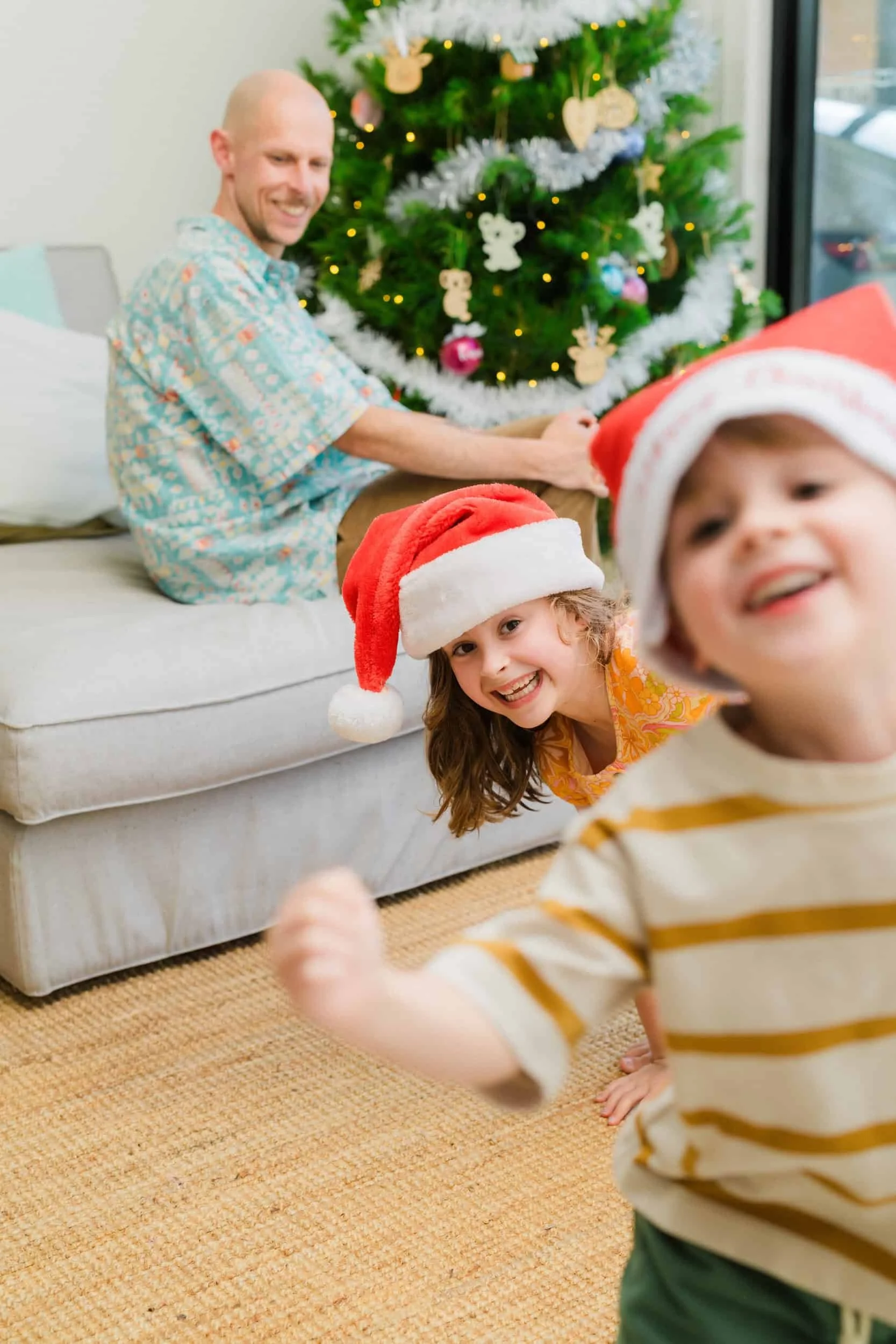 A man and two children wearing Santa hats in front of a decorated Christmas tree, celebrating the holidays.