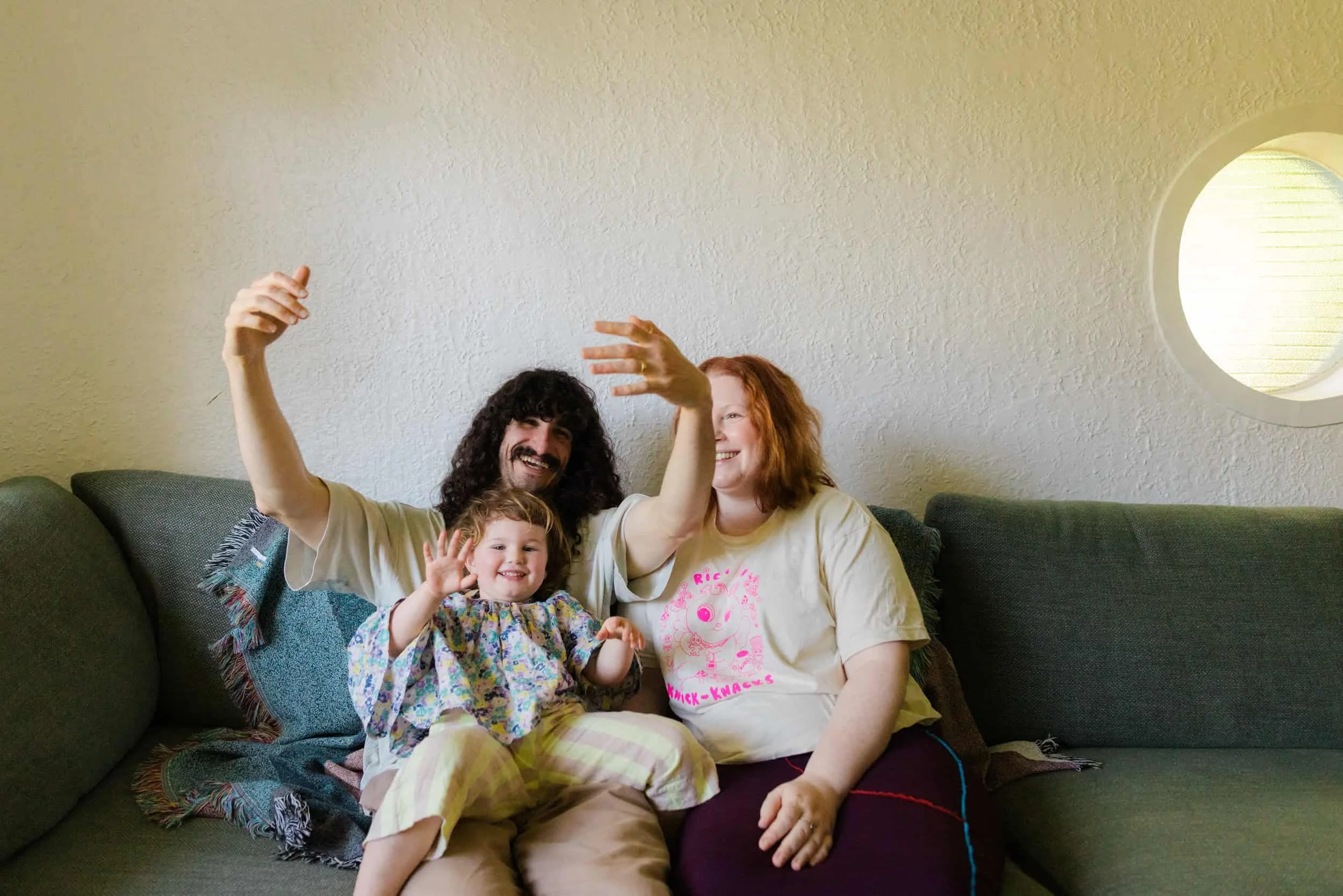 A happy family sitting on a green sofa, waving and smiling at the camera. The family consists of a man, a woman, and a young girl, in a well-lit room with a round window in the background.