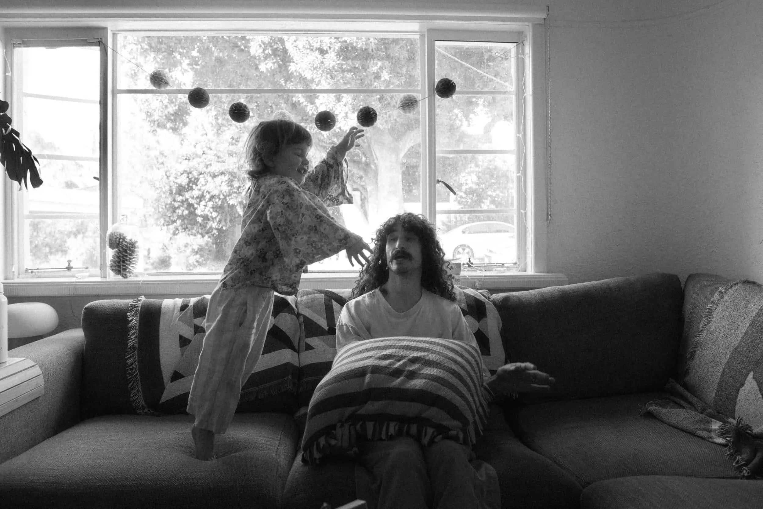 A black and white photo of two children playingly interacting indoors. One child is sitting on a couch with a pillow on their lap, while the other child stands on the couch, reaching out towards the seated child's head. The background shows a large w