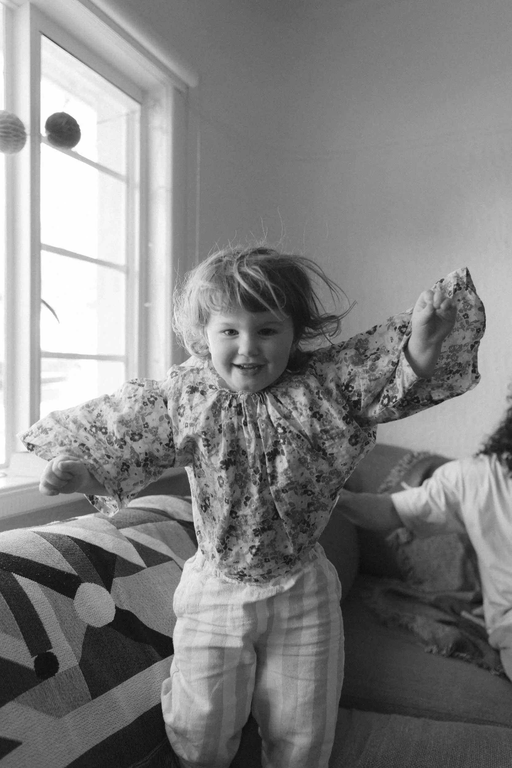 Young girl with curly hair in a floral blouse and checkered pants jumping onto a sofa, smiling, in a bright living room with a window.