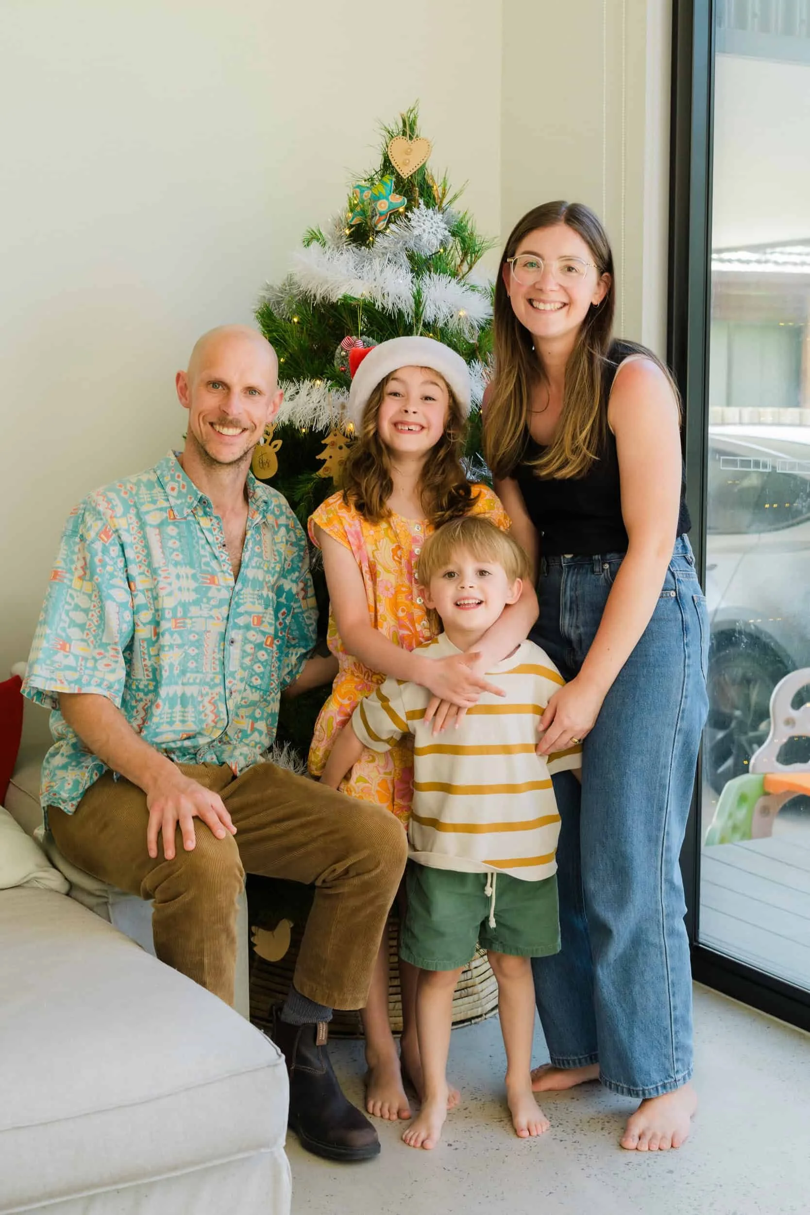 Family of four celebrating Christmas in front of a decorated Christmas tree indoors, with the mother and daughter smiling and wearing holiday attire, and the father and son sitting on the floor.
