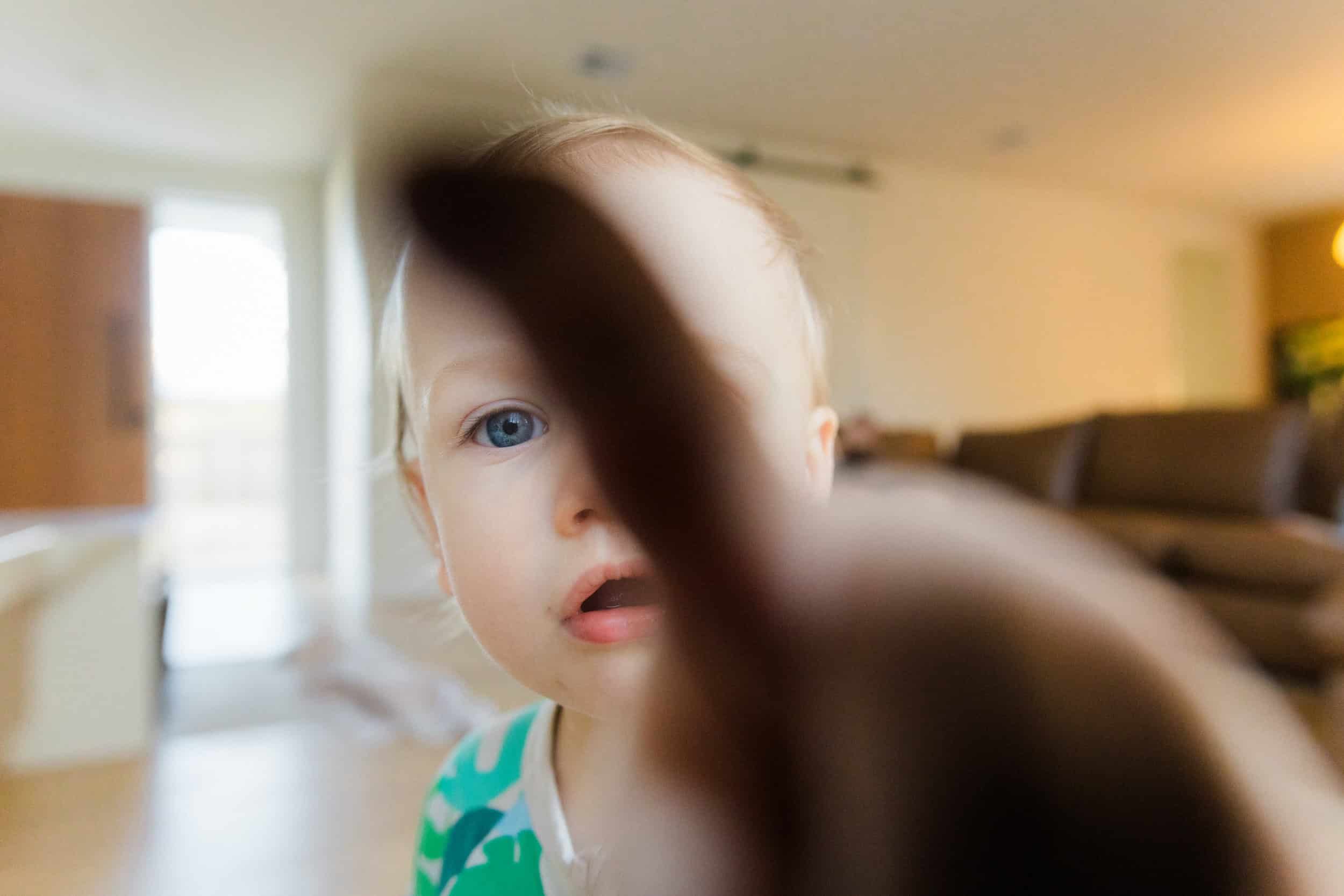 Close-up of a young child with blue eyes and blonde hair, reaching towards the camera with a finger, in a bright, indoor setting with blurred background.