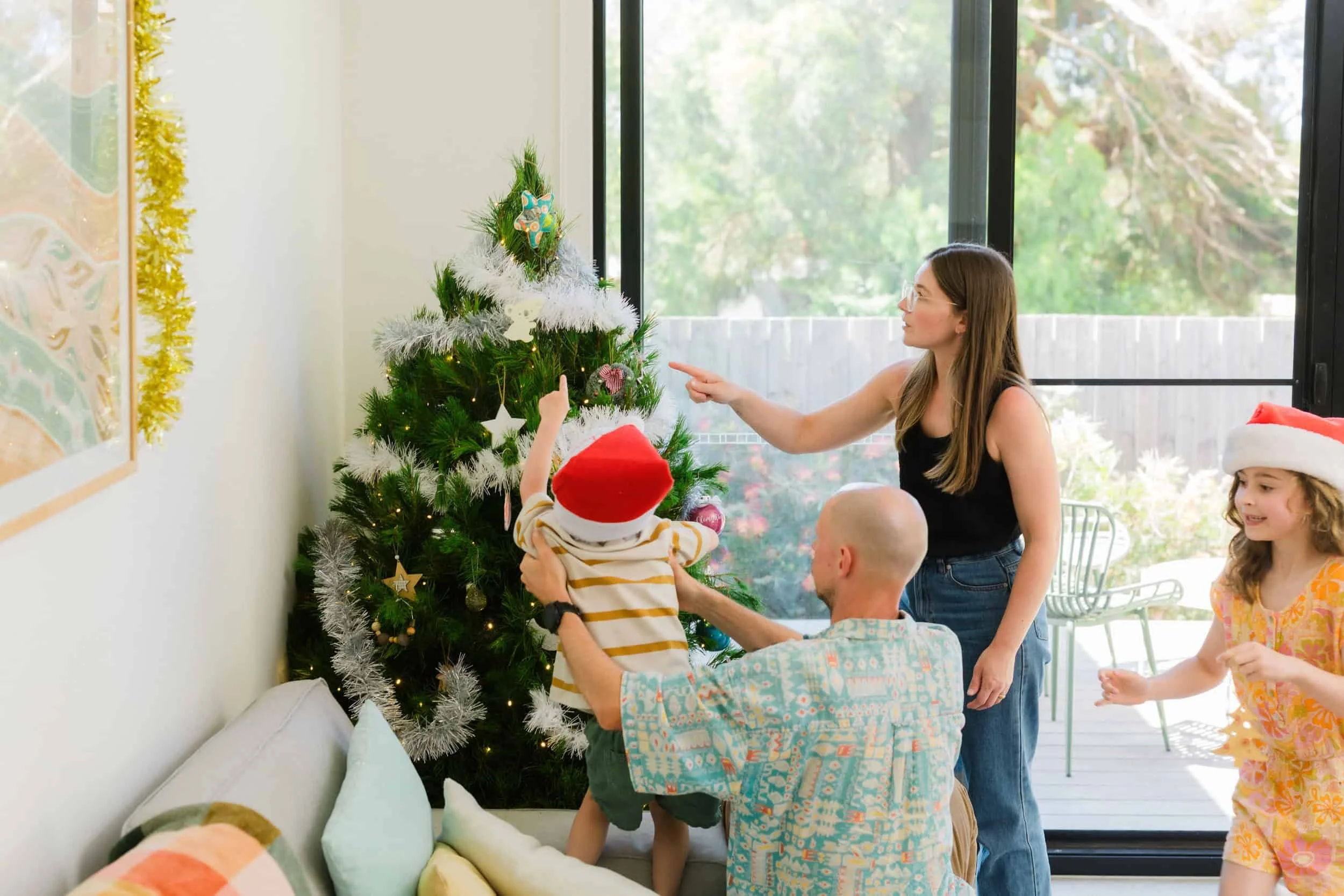 A family decorating a Christmas tree in a bright room with large windows. The woman is pointing at the tree, and a man is holding a young child wearing a Santa hat, reaching towards the tree. A young girl in a Santa hat and summer dress is nearby, sm
