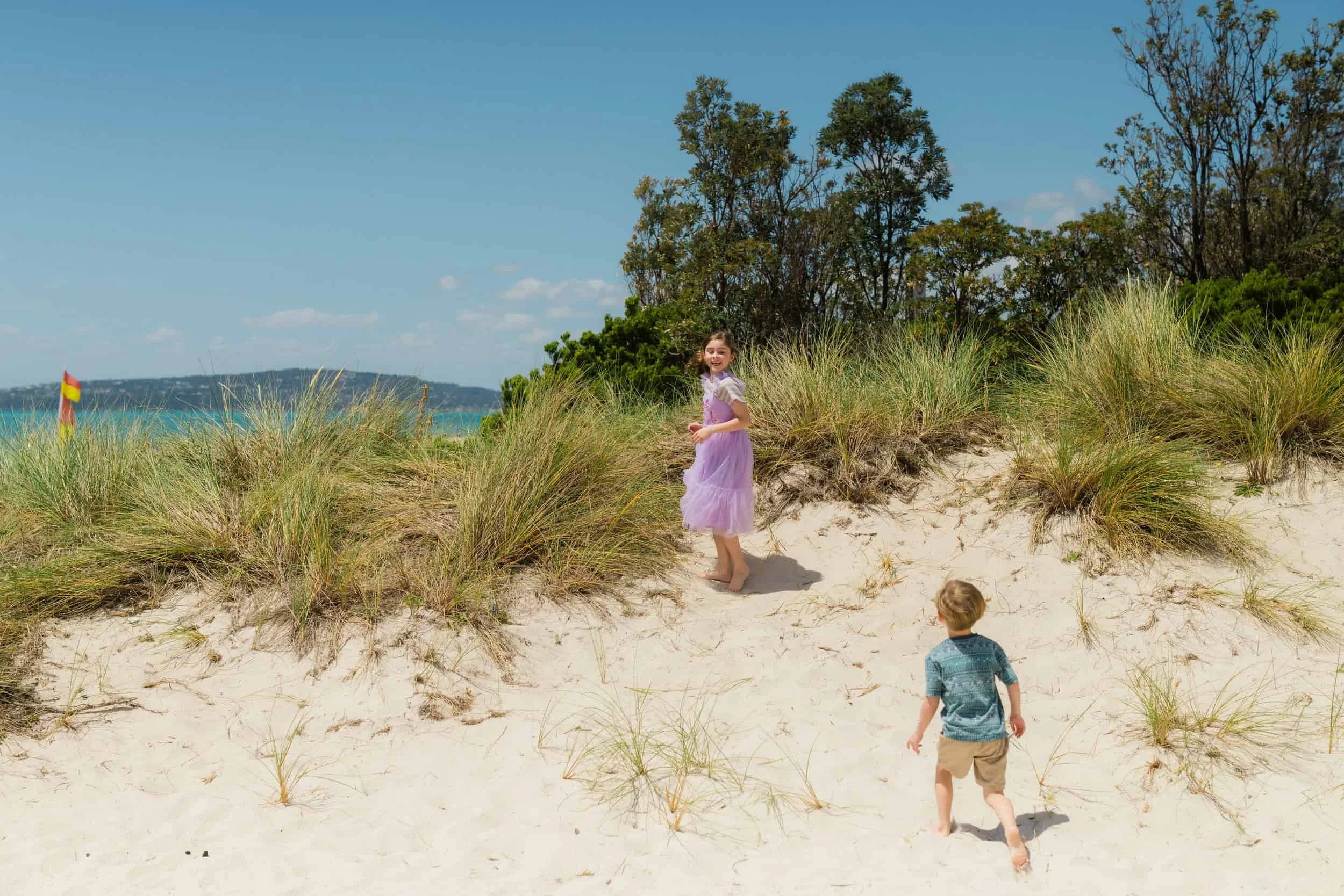 Two children, a girl in a purple dress and a boy in a blue shirt and khaki shorts, playing on sandy dunes with green beach grass and trees behind them under a blue sky with clouds.