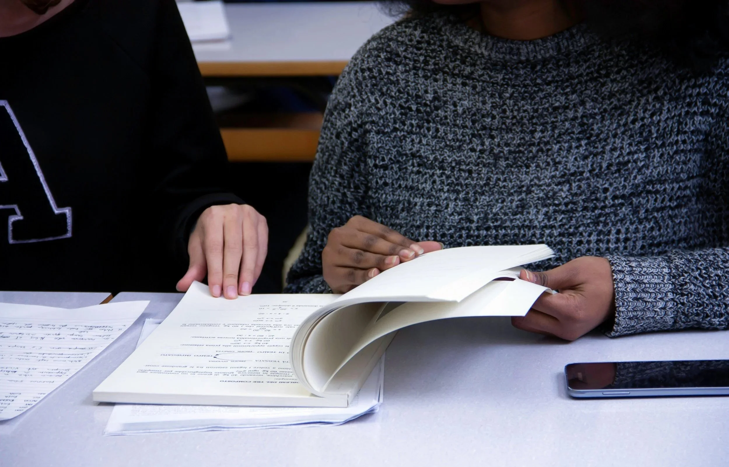 Two people sitting at a table, one flipping through a book, with papers and a mobile phone on the table.