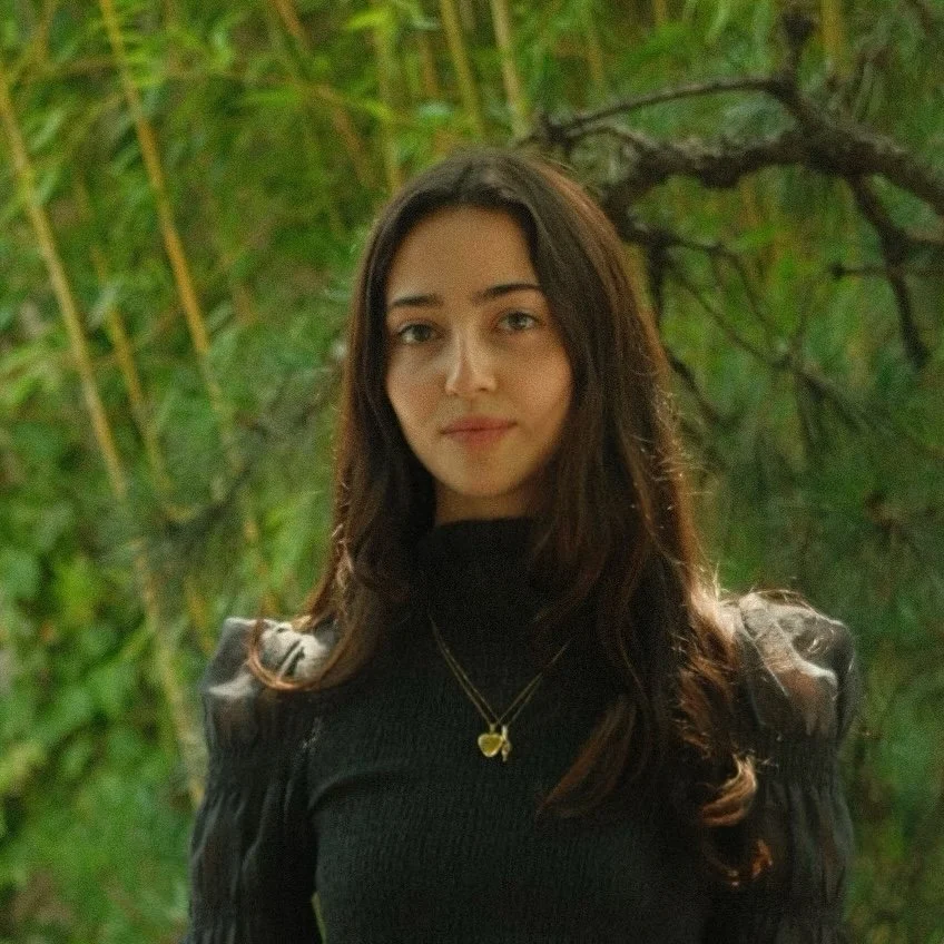 A young woman with long dark hair, wearing a black top and a butterfly necklace, standing outdoors in front of green foliage and trees.
