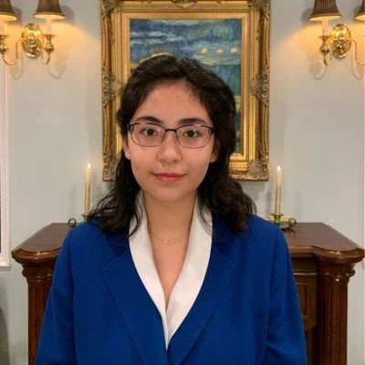 A young woman with glasses and shoulder-length dark hair standing indoors in front of a painting with candles and wall sconces, wearing a blue blazer over a white shirt.