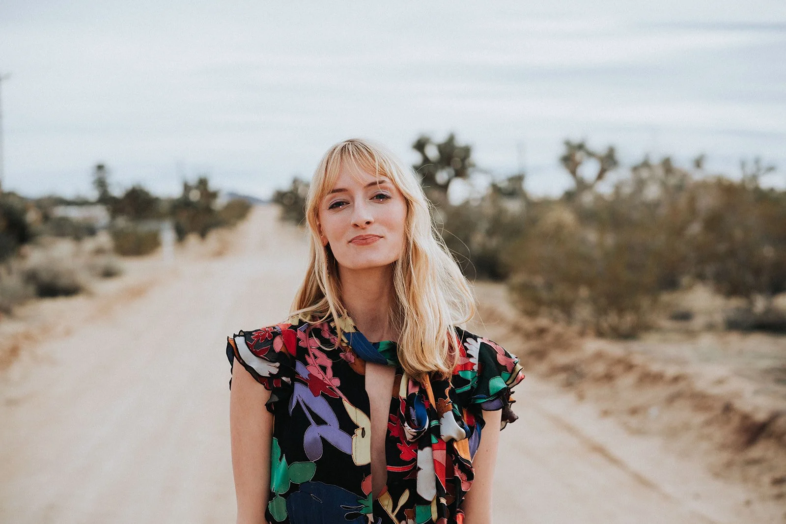 A woman with blonde hair in a colorful floral dress standing on a dirt path in a desert landscape with sparse vegetation and a cloudy sky.