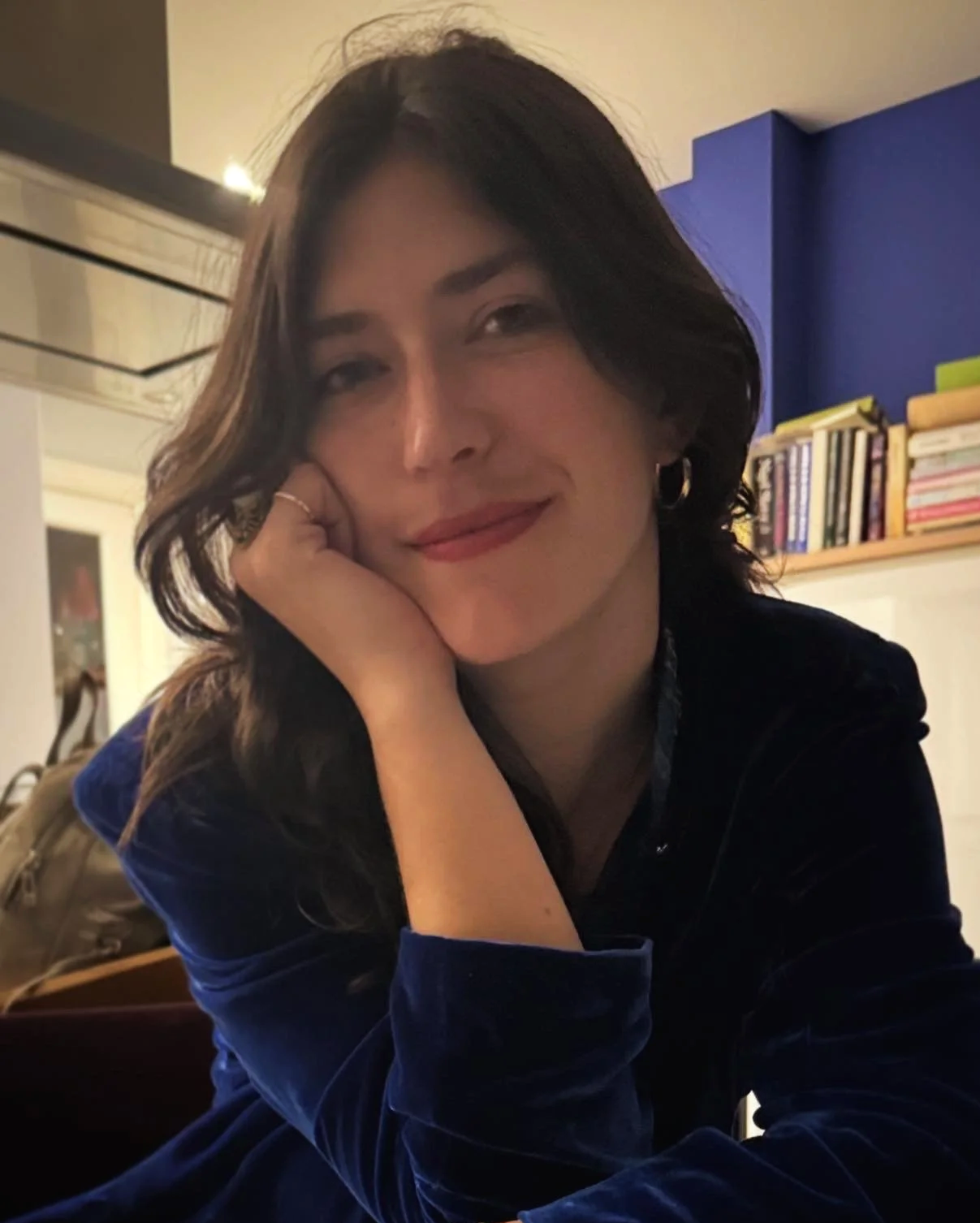 A woman with dark brown hair, wearing a dark blue velvet top, resting her head on her hand and smiling at the camera in a room with bookshelves and a mirror in the background.