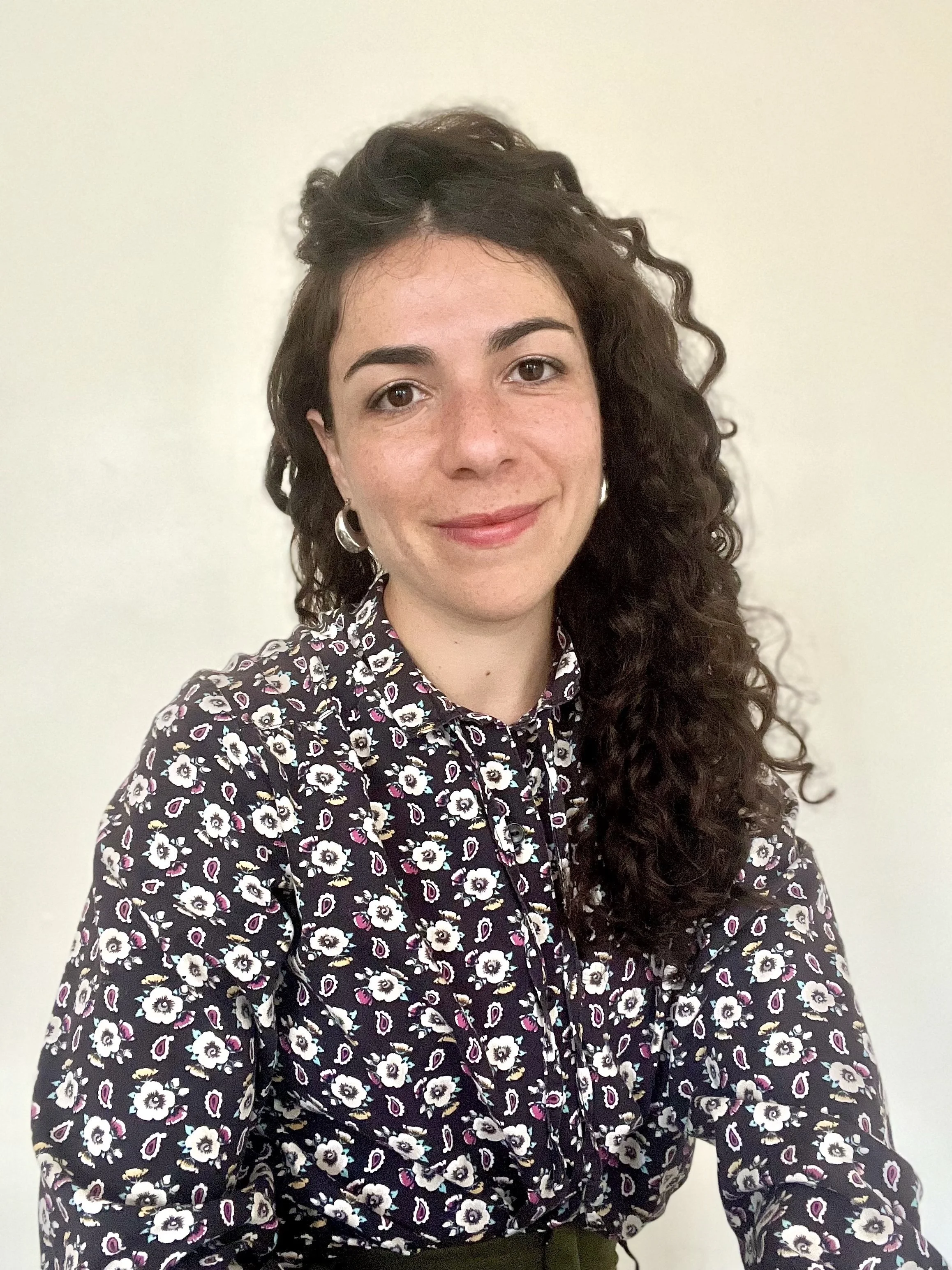A woman with curly dark hair wearing a floral patterned blouse, smiling, in front of a plain white wall.