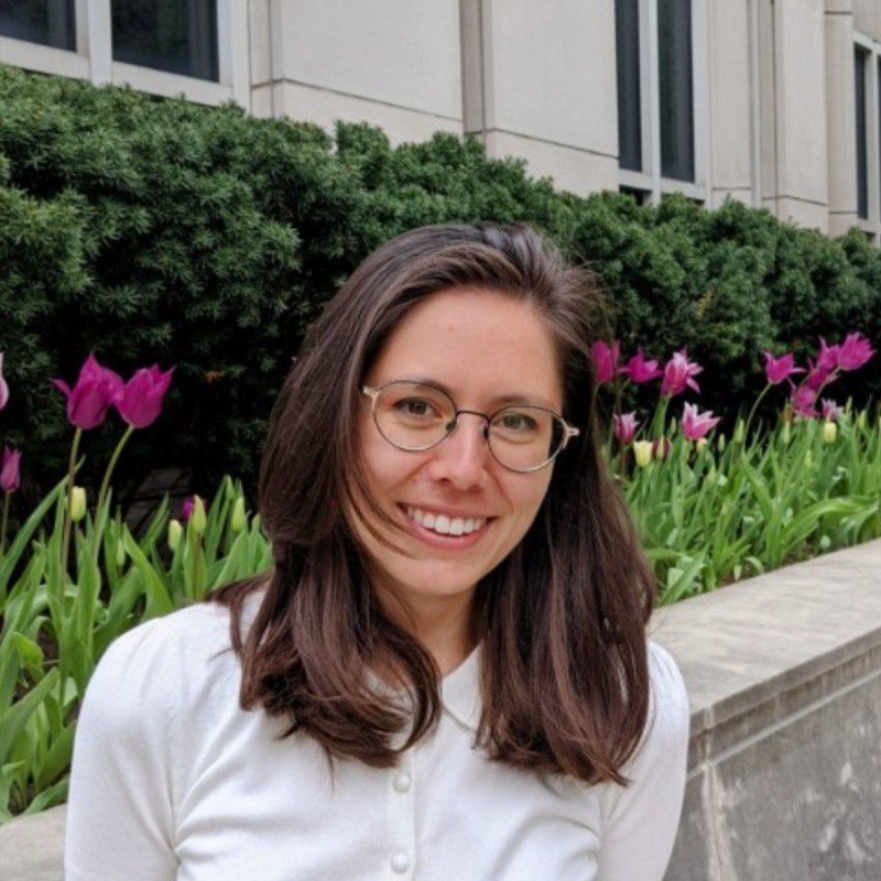 A young woman with shoulder-length brown hair, glasses, and wearing a white shirt, smiling outdoors with purple tulips and green bushes in the background.