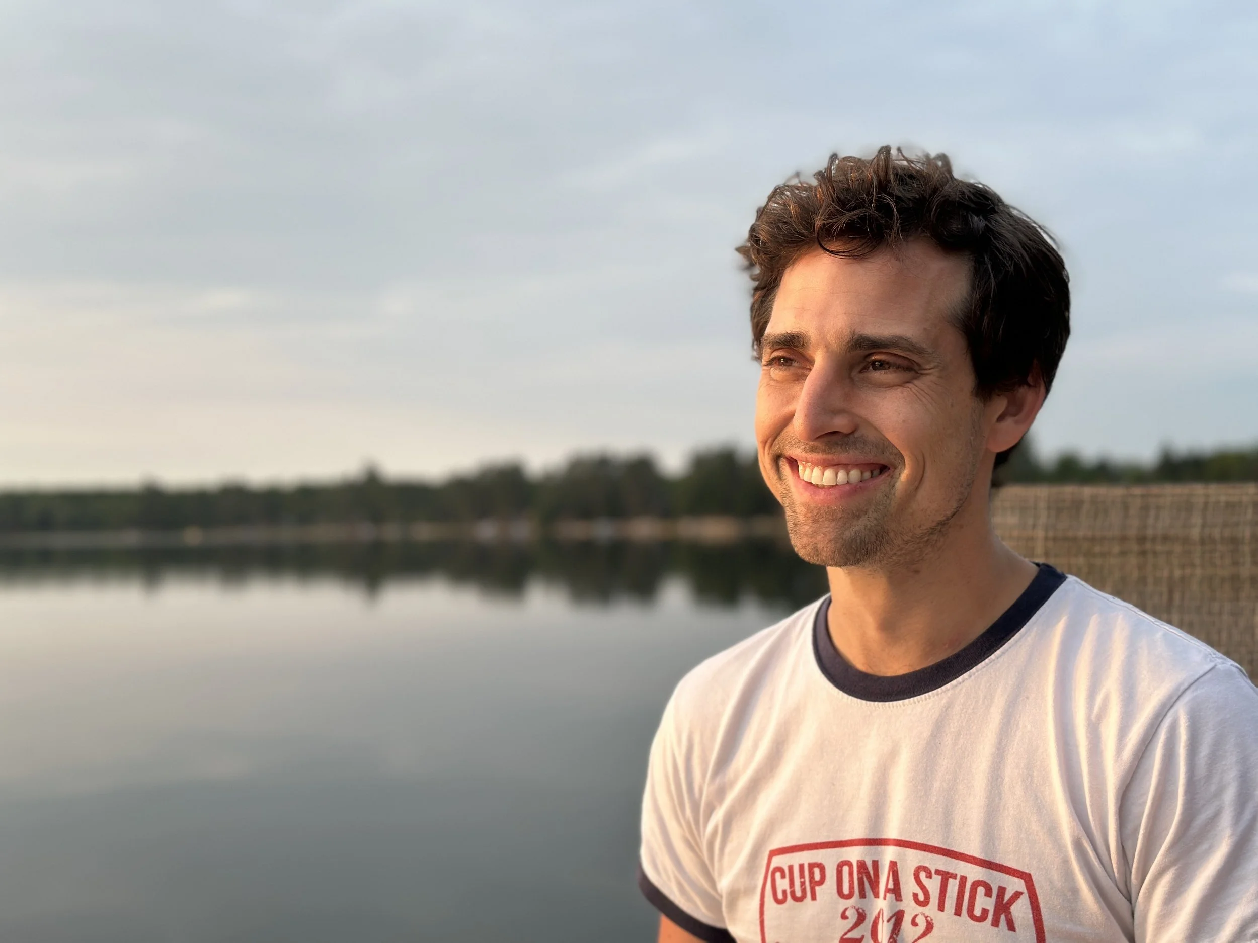 A smiling man with dark, wavy hair looking to the right, standing by a calm body of water with a distant tree-lined shoreline under a cloudy sky.