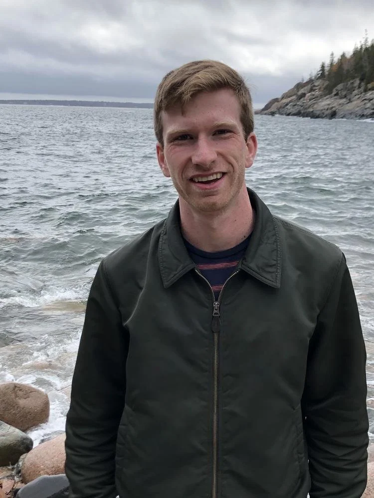 A young man with short hair and a big smile standing on a rocky beach near the water, with a cloudy sky and distant trees along the shoreline in the background.