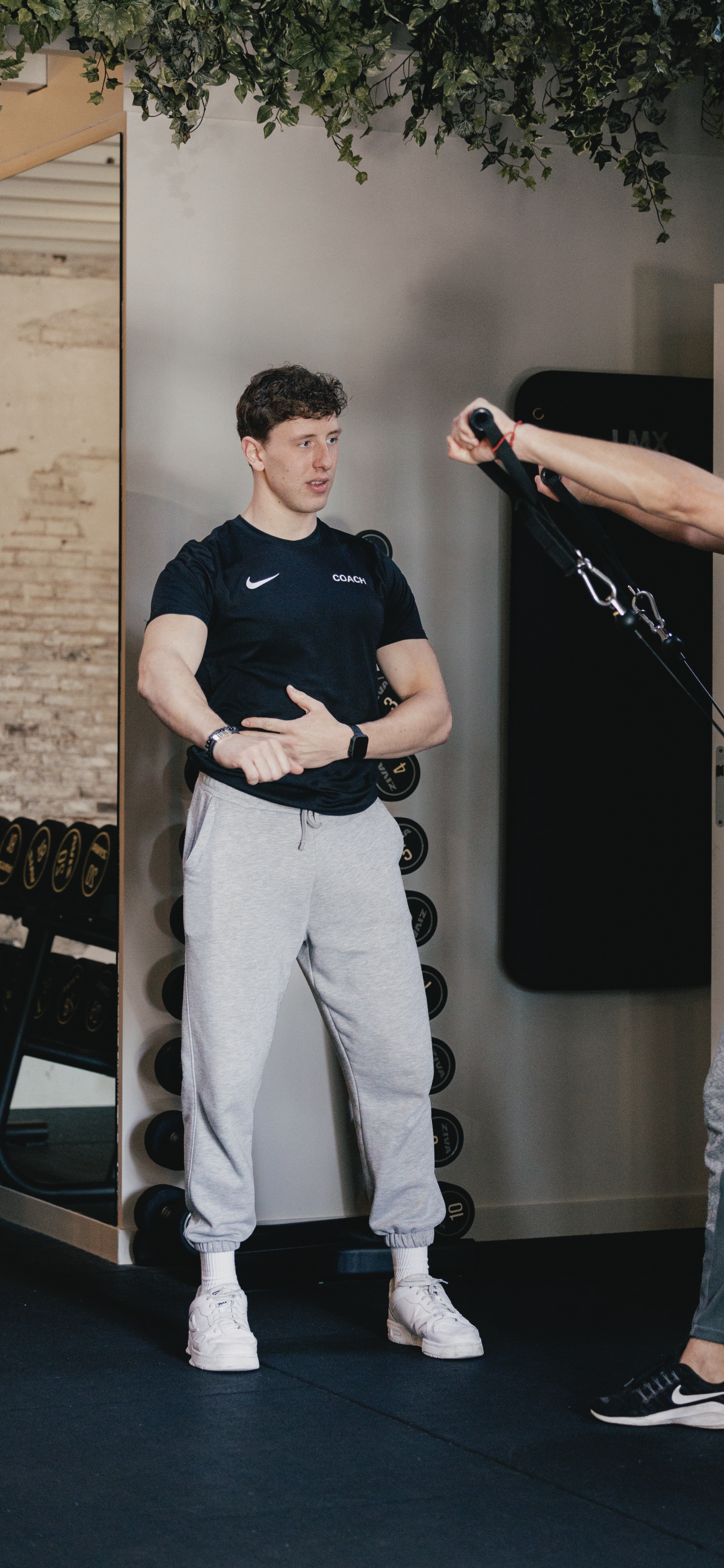 Persoon in sportsoutfit met 'Coach' op shirt, in een sportschool in gesprek met trainer die apparaten gebruikt.